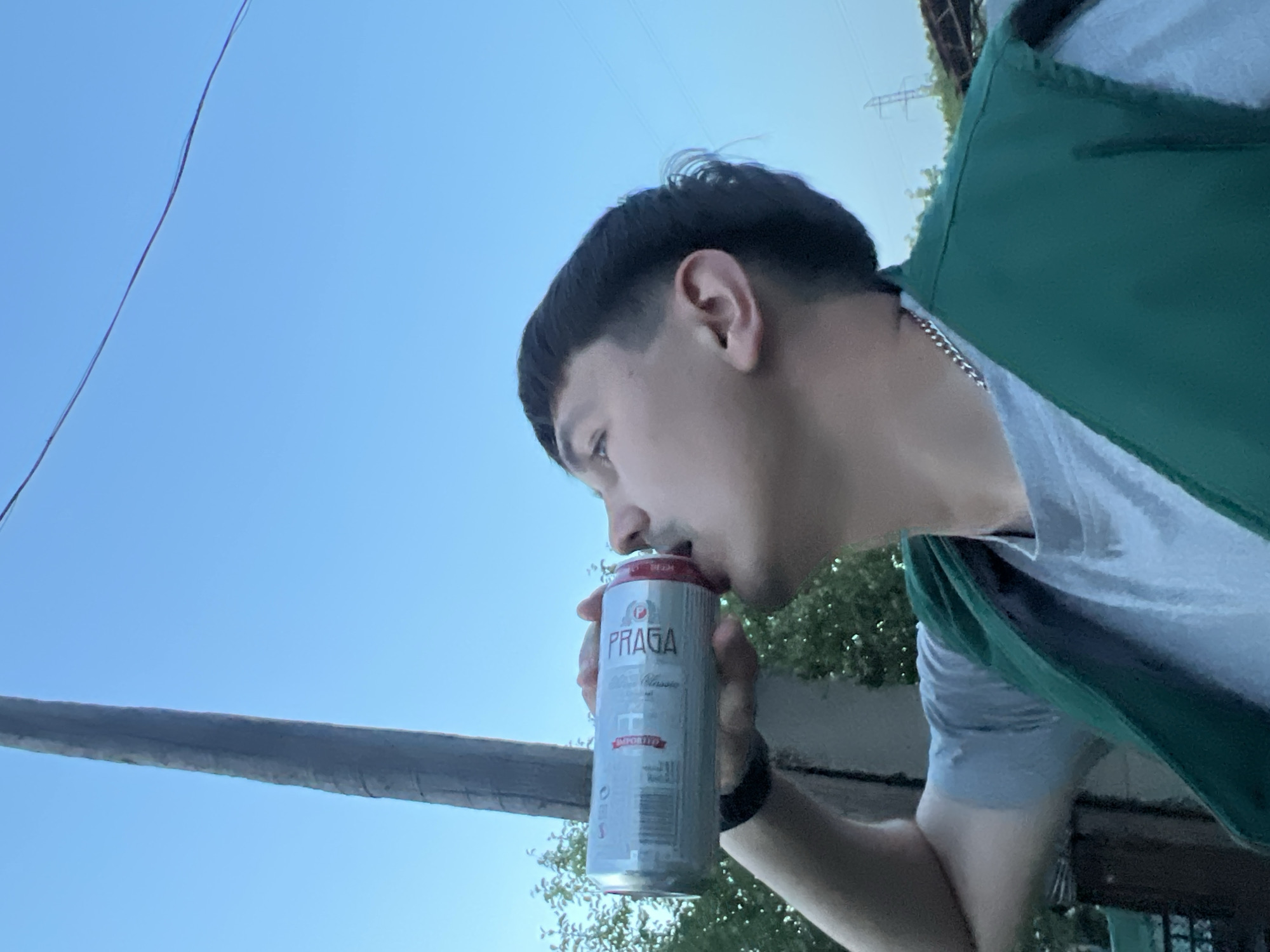 A person is drinking from a silver can of Praga beer in an outdoor location. The person has dark hair and is wearing a grey t-shirt and a green vest. A grey metal bar is to the left of the can. In the background, there is a blue sky, some green trees, and power lines. Based on the brand of beer and other details, it is likely taken in a location where Praga beer is available, possibly in Chaikovsky, Russia.
