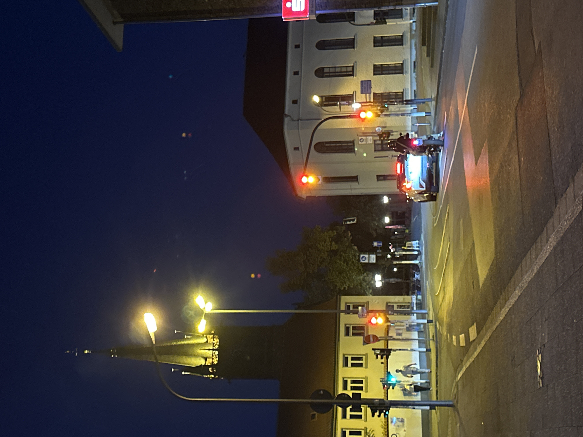 The image depicts a street scene at night in Krefeld, Germany, likely near a church with a prominent spire. A white building with arched windows stands to the right, and a smaller, lighter colored building with rectangular windows is visible to the left. Traffic lights are illuminated, showing red and yellow signals. A white car with its brake lights on is stopped at an intersection, facing away from the camera. A motorcycle with a rider is positioned behind the car. Pedestrians are visible walking on the sidewalk in front of the building on the left. Streetlights illuminate the road and sidewalks, casting yellow and white light. The sky is dark blue, indicating nighttime.