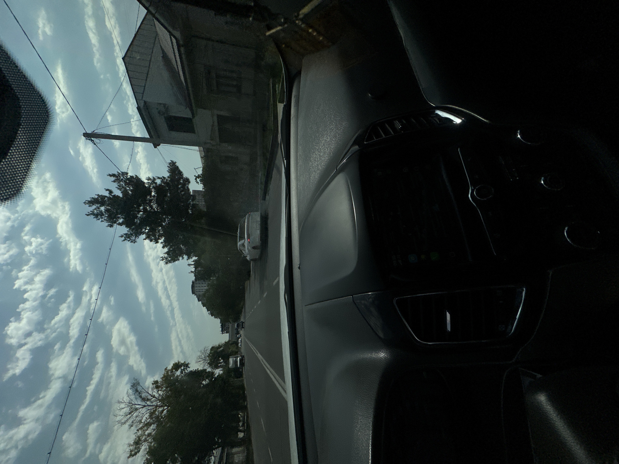 The image, taken from inside a vehicle, captures a view of a city street in Tashkent, Uzbekistan. The foreground is dominated by the dark interior of the car, including the dashboard and steering wheel. Beyond the car's windshield, a tree-lined street is visible, with several cars driving in both directions. In the background, residential buildings and more trees can be seen. The sky is partly cloudy, with distinct cloud formations creating a layered effect. Power lines and a utility pole are also present in the scene. The lighting suggests it is daytime, possibly late morning or early afternoon.