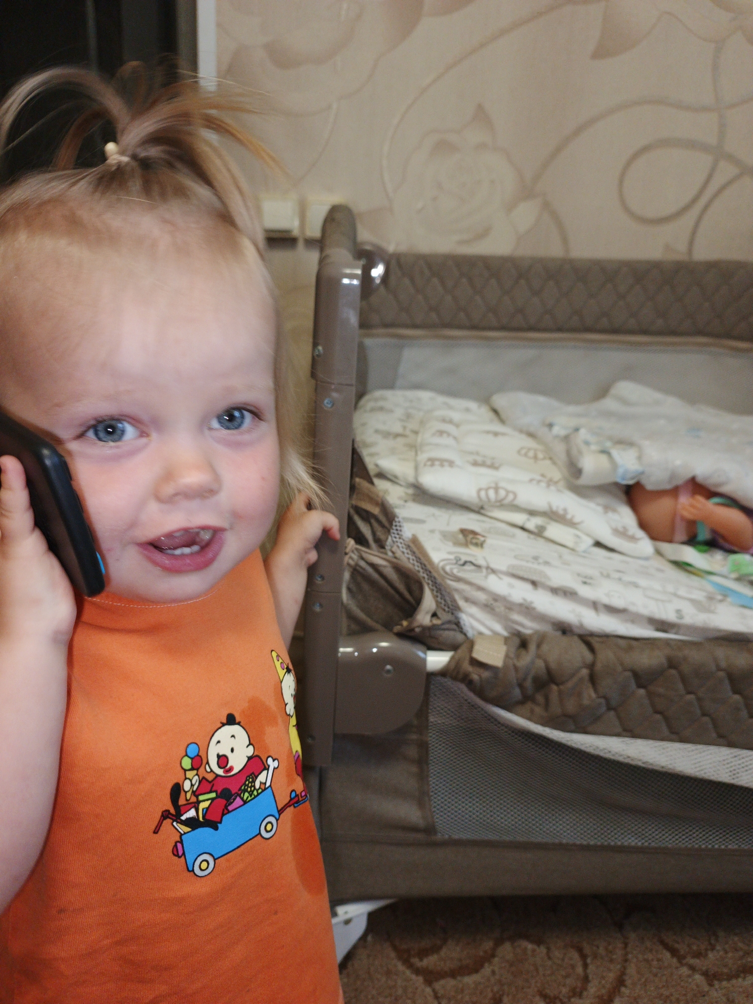 An undated, eye-level, medium shot shows a young Caucasian child with blonde hair in a ponytail, holding a dark-colored mobile phone to their left ear. The child is wearing an orange t-shirt with a cartoon illustration of a child in a blue wagon holding an ice cream cone. The child is looking at the camera with wide blue eyes and an open mouth, appearing to be smiling. The child's right hand is visible grasping the side of a brown-colored crib. The crib is filled with light-colored bedding, and a partially visible doll can be seen lying on its back. The background of the image shows a wall with patterned wallpaper and a portion of a wooden closet door. The scene appears to be indoors, possibly in a bedroom or nursery. There is no specific indication of the location within the city of Zlatoust, Russia.