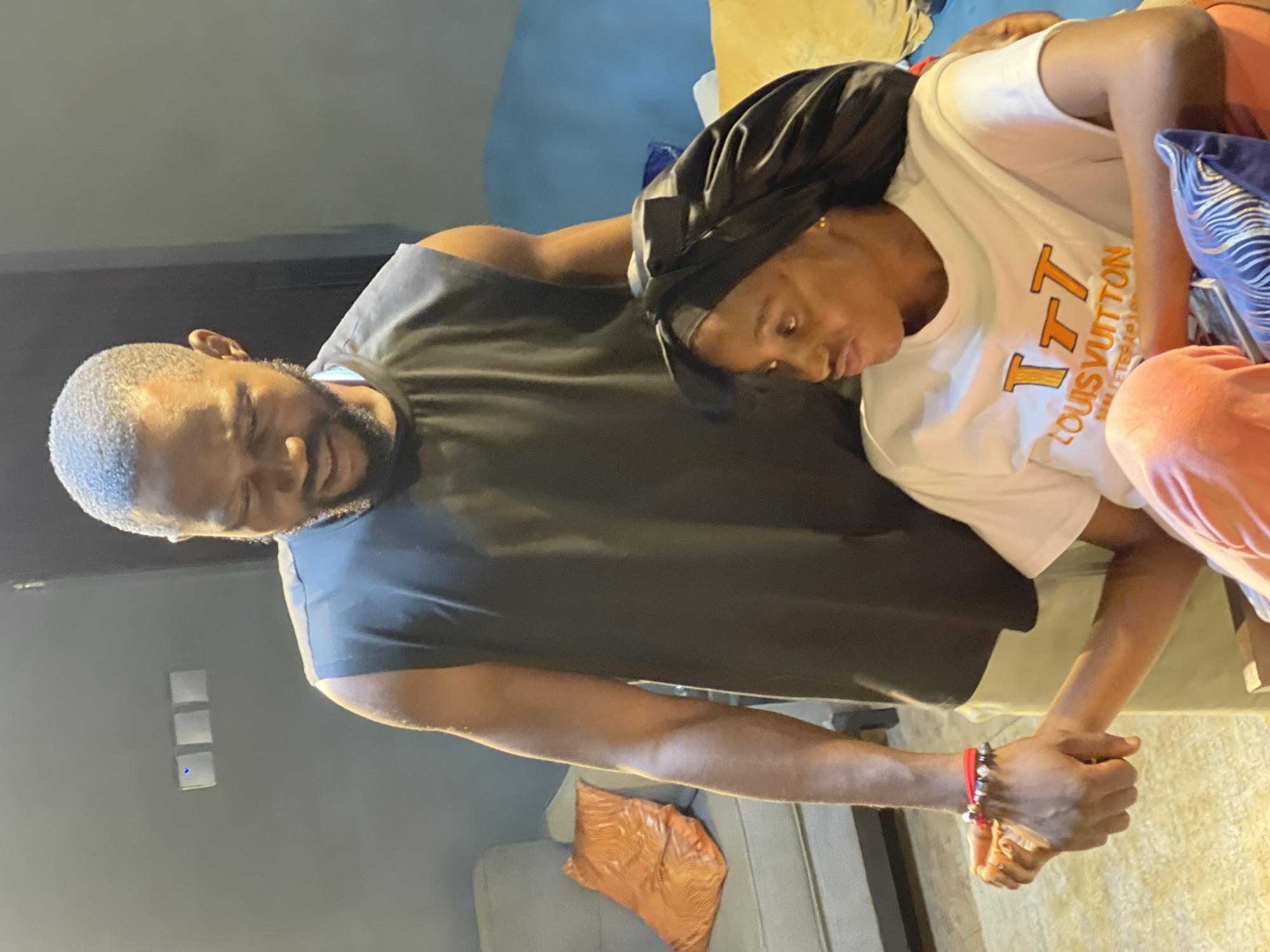 A man and a woman are indoors, appearing to be in a living room. The man, with a beard and short hair, is wearing a black sleeveless shirt and tan pants. He is standing and looking down at the woman. The woman is seated, wearing a white t-shirt with an orange Louis Vuitton logo and orange pants. She is also wearing a black satin bonnet. They are holding hands, with the man's hand over hers. The room has light-colored walls and furniture, including a gray sofa with an orange pillow and a blue sofa. There are light switches on the wall. The overall lighting suggests it is daytime or an indoor setting with artificial light. The location is identified as Abuja, Nigeria.