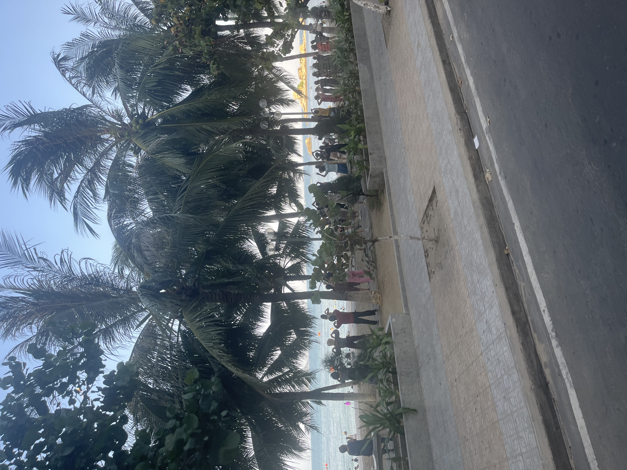 The image depicts a lively beach scene in Nha Trang, Vietnam, with a group of people engaged in what appears to be a synchronized exercise routine on the sand. Palm trees frame the view, their fronds swaying gently against a clear sky. The ocean stretches out in the background, dotted with a few swimmers and what might be small boats. A paved walkway runs along the edge of the beach, separating it from a street. The overall atmosphere is one of a relaxed, tropical resort town.