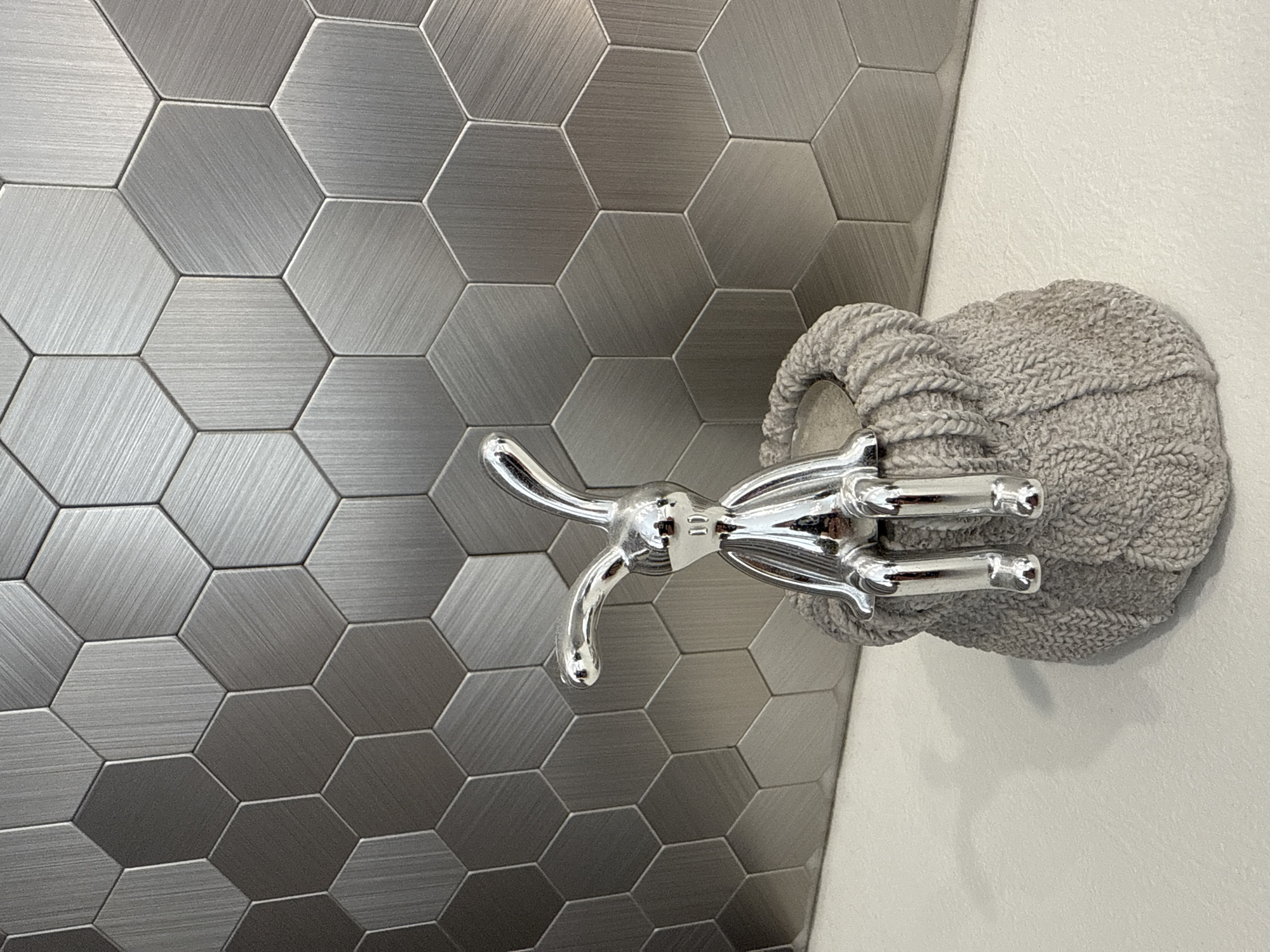 A close-up, rotated view shows a gray knitted pot holder mounted on a white wall, next to a gray hexagonal tiled wall. A chrome metal faucet fixture is attached to the pot holder. The location is not determinable from the image.