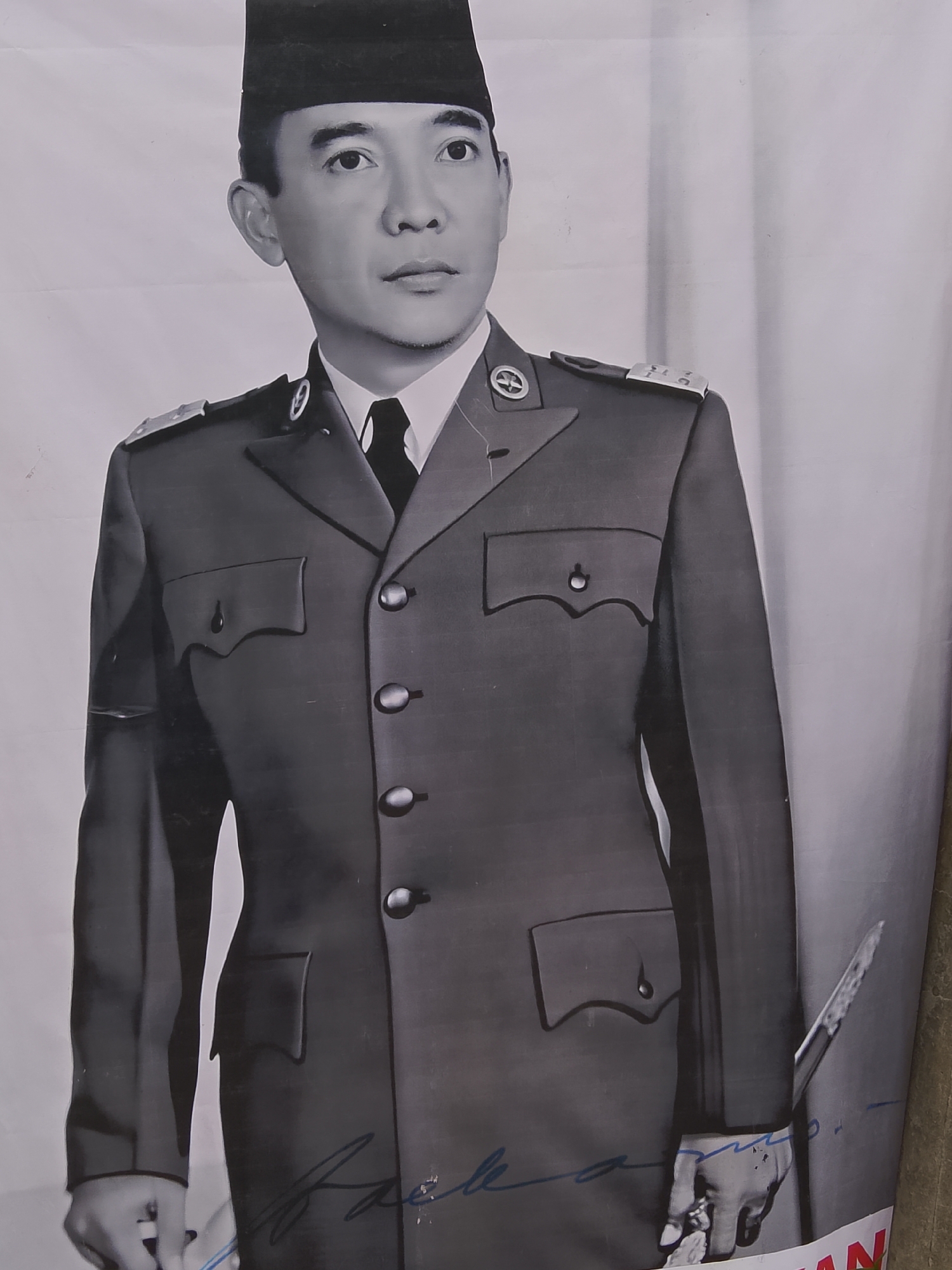 A black and white, medium shot portrait shows a man wearing a dark military-style uniform with a black cap. The man faces forward with a neutral expression. He has dark hair and is wearing a white collared shirt with a dark tie. The uniform has epaulets with insignia on the shoulders, and buttons down the front. There are pockets on the chest and abdomen of the jacket. The man's right hand is visible at the bottom right of the frame, holding a dagger. A blue signature is visible on the lower part of the image. The background is a plain, light-colored surface, likely a poster. The man's identity is Sukarno, the first President of Indonesia. The location is stated to be in the city of Cikukulu, Indonesia.