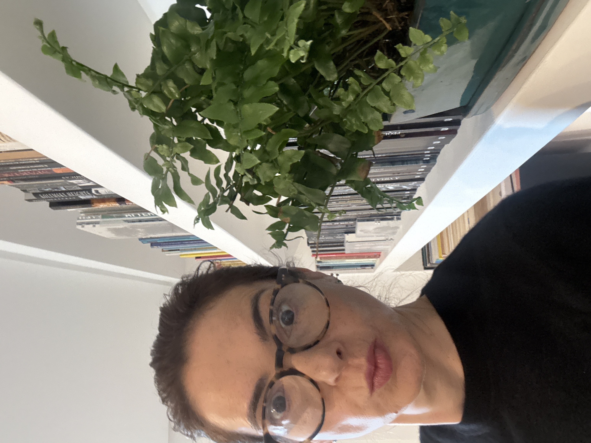 A woman with glasses is looking at the camera. She is standing near a bookshelf with books and a potted plant. The location is impossible to determine from the image.
