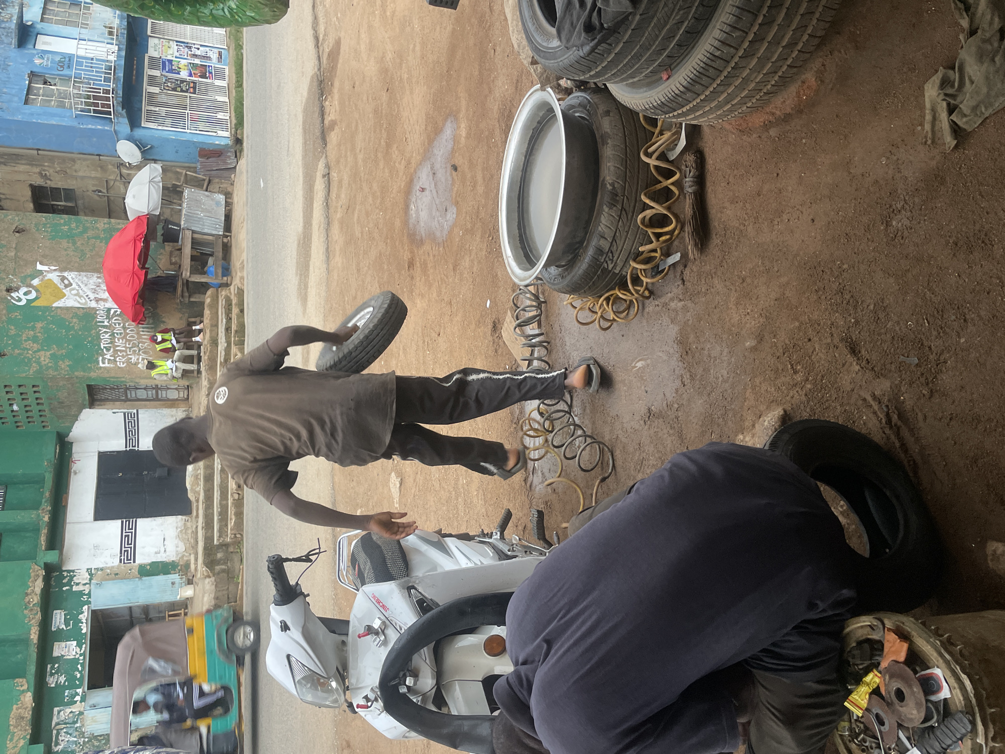 This image depicts an outdoor tire repair and service scene in Jos, Nigeria, during the daytime. The ground is dusty brown earth.
In the foreground, two men are engaged in work. To the left, a man in a brown shirt and dark pants, wearing sandals, walks while holding a small tire. In the lower right, another man, dressed in a dark blue top and dark pants, is bent over a white JINCHENG brand scooter, seemingly working on its tire or a related component. Beside him, a metal container holds various small tools and tire patches.
Numerous tires are present: some are stacked, one is being held, and a large car tire with a silver rim filled with water, likely for puncture detection, sits on the ground. Coiled yellow air hoses are visible near the tires, along with a small broom.
In the mid-ground, a road runs across the image. On the road, a yellow and green three-wheeled auto-rickshaw is visible. Beyond the road, several buildings line the street. A prominent blue building features "NYSC" and "Goby" text, along with a satellite dish. Next to it, a green-painted building has a sign that partially reads "FACTORY WORKERS NEEDED 55,000 708." Underneath red and white umbrellas, several people are seated, some wearing yellow/green reflective vests, possibly customers or employees taking a break. The scene portrays a typical bustling roadside service shop.