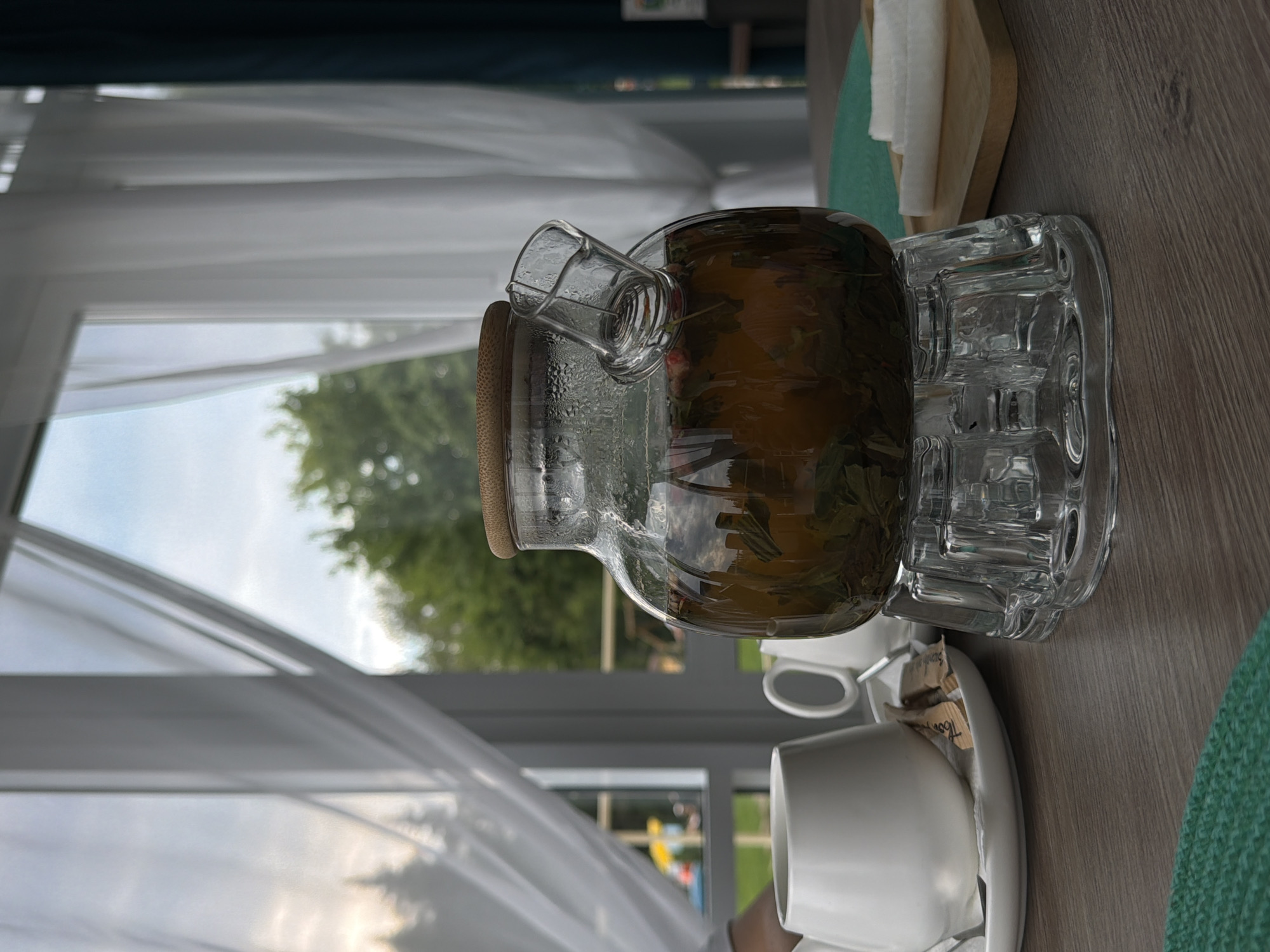 The image shows a glass teapot filled with herbal tea, sitting on a clear glass stand. The teapot has a wooden lid and a small glass spout. In front of the teapot, there is a white ceramic pitcher and two white ceramic cups on a saucer. There are also two small packets of sugar on the saucer. In the background, there is a window with white curtains, through which trees and a cloudy sky can be seen. The scene is set on a wooden table. There are no people visible in the image. This scene depicts a relaxing moment, likely in a cafe or restaurant, in the city of Chusovoy, Russia.