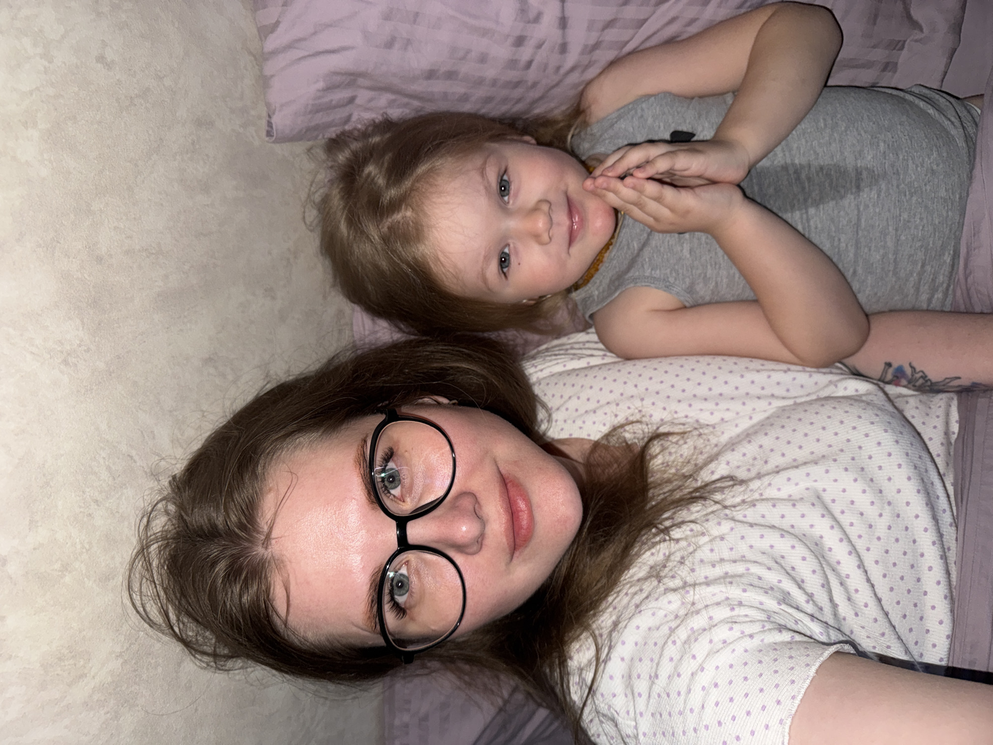 The image shows a selfie of a woman with long brown hair and round black glasses, lying down next to a young child with blonde hair and blue eyes. The woman is wearing a white pajama top with small purple polka dots. The child is wearing a gray t-shirt. They are both lying on a bed with lavender-colored linens and pillows. The background includes a wall with a light beige textured surface. The setting appears to be indoors, possibly a bedroom. This image does not provide specific details that would allow one to ascertain the location as Gornyi (Dvurechenskoe S/P), Russia.