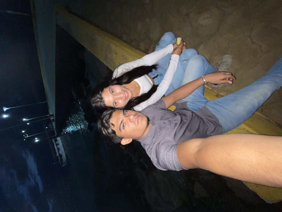 A young couple poses for a selfie at night near the water. The woman, wearing a white long-sleeved top and jeans, sits next to the man in a dark button-down shirt and jeans. They're positioned near a yellow ledge, with the water visible in the background along with street lights. The man's arm is outstretched to take the photo.