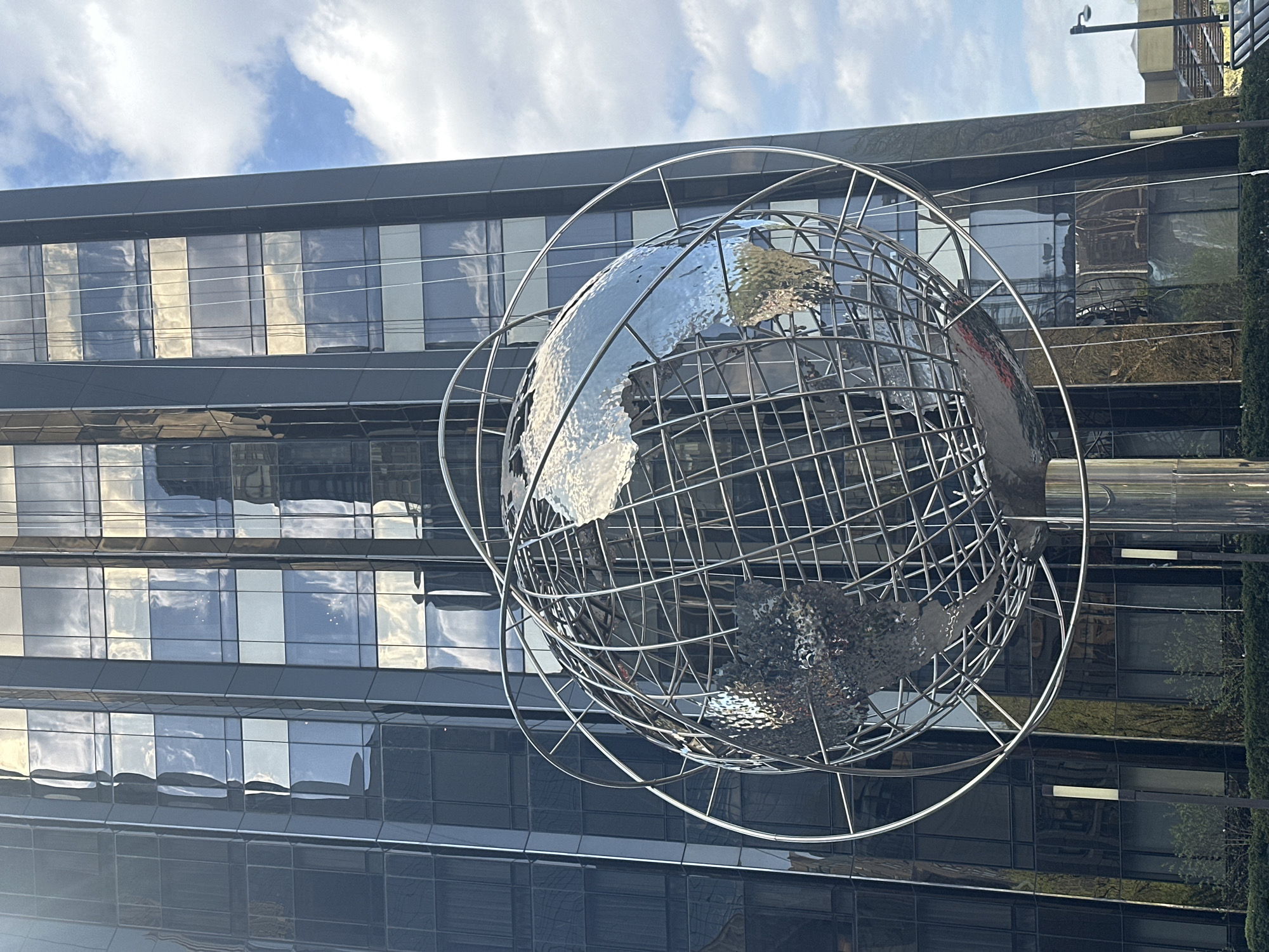 The image captures a view of the Columbus Circle Globe, a prominent public art installation located in Columbus Circle in New York City. The globe is depicted as a shiny, metal structure representing the Earth, enclosed within a framework of intersecting metal rings. It stands in front of a modern building with a dark, reflective facade composed of numerous window panes. The windows reflect the sky, clouds, and surrounding environment. To the right of the globe, a vertical strip of greenery suggests nearby landscaping or trees. The sky is visible in the upper portion of the image, with light clouds scattered across it. The image appears to be taken from a relatively low angle, looking upward at the globe and the building.