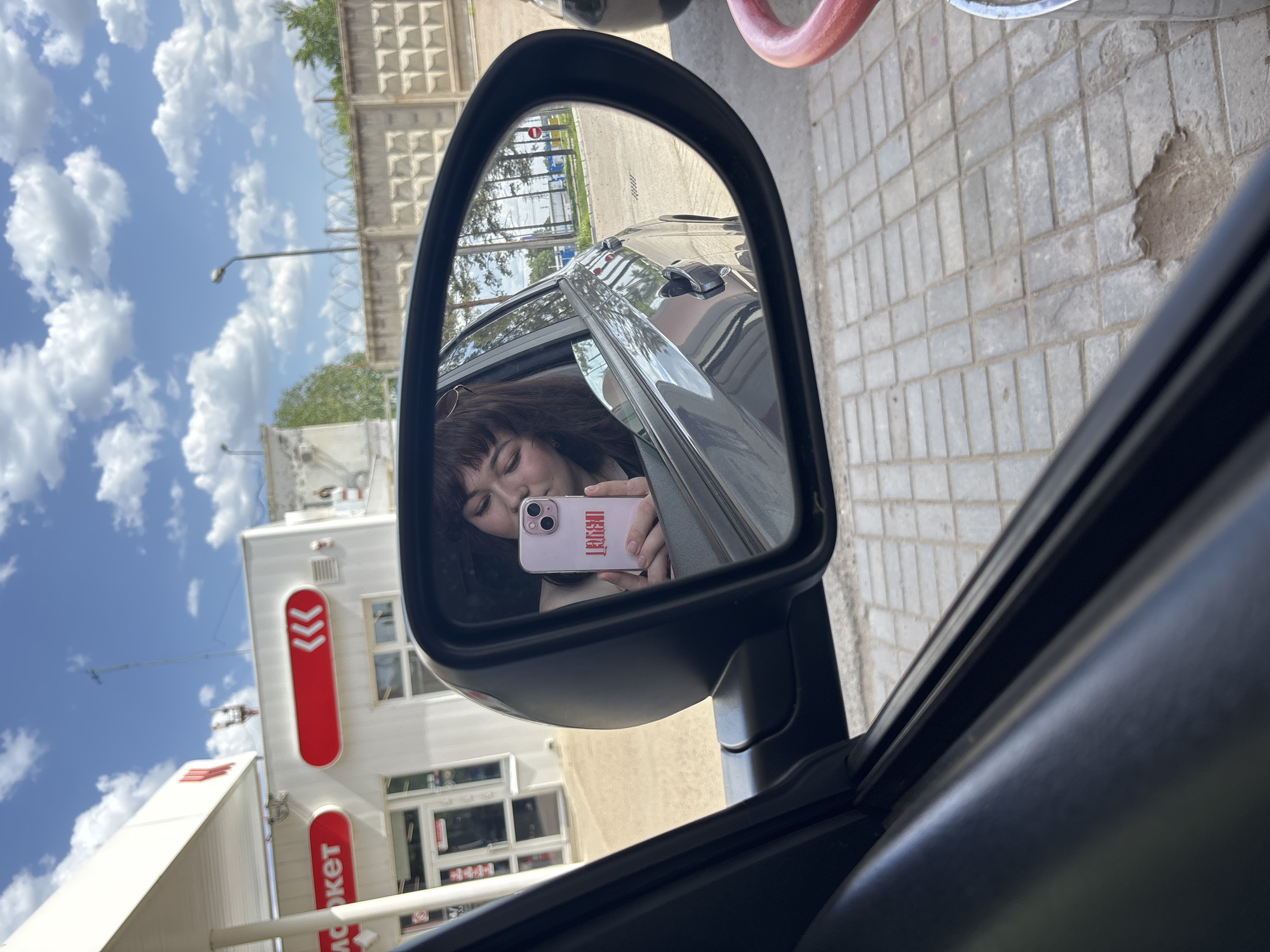 A rearview mirror reflects a young woman taking a selfie with her phone. The background shows the exterior of a Lukoil gas station in the city of Chaikovsky, Russia, as indicated by the red and white Lukoil logo and branding. The gas station building is white with multiple windows and a red awning. The sky is blue with scattered clouds. The mirror also reflects the car's interior, including part of the door. The person holding the camera is in the car.