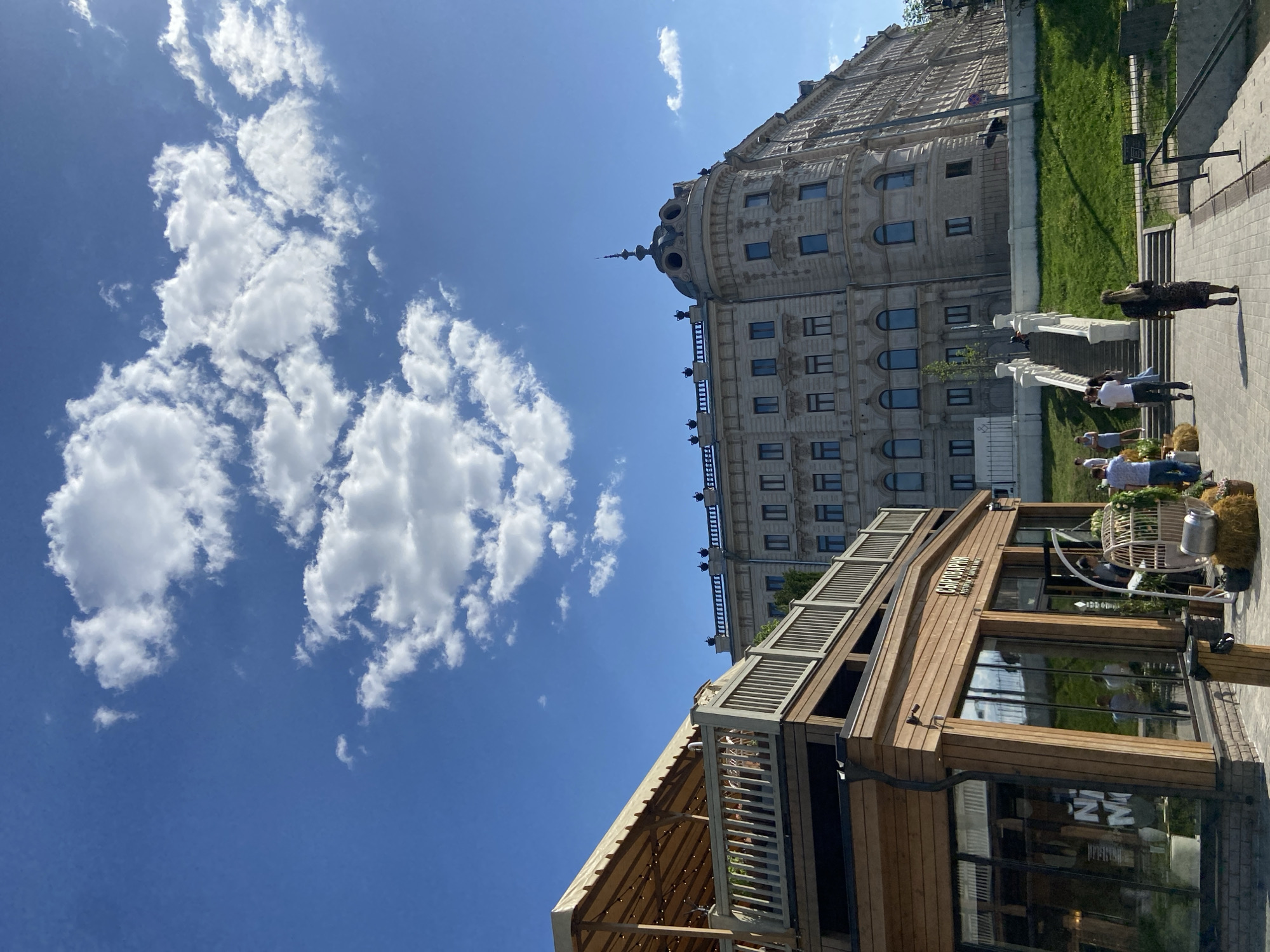 The image depicts a sunny daytime scene in Kazan, Russia. In the foreground, a wooden structure with a partially covered balcony is visible. Behind it is a large, light-colored building with multiple windows and ornate architectural details. People are present on a pathway near the large building. The sky is blue with scattered puffy white clouds.