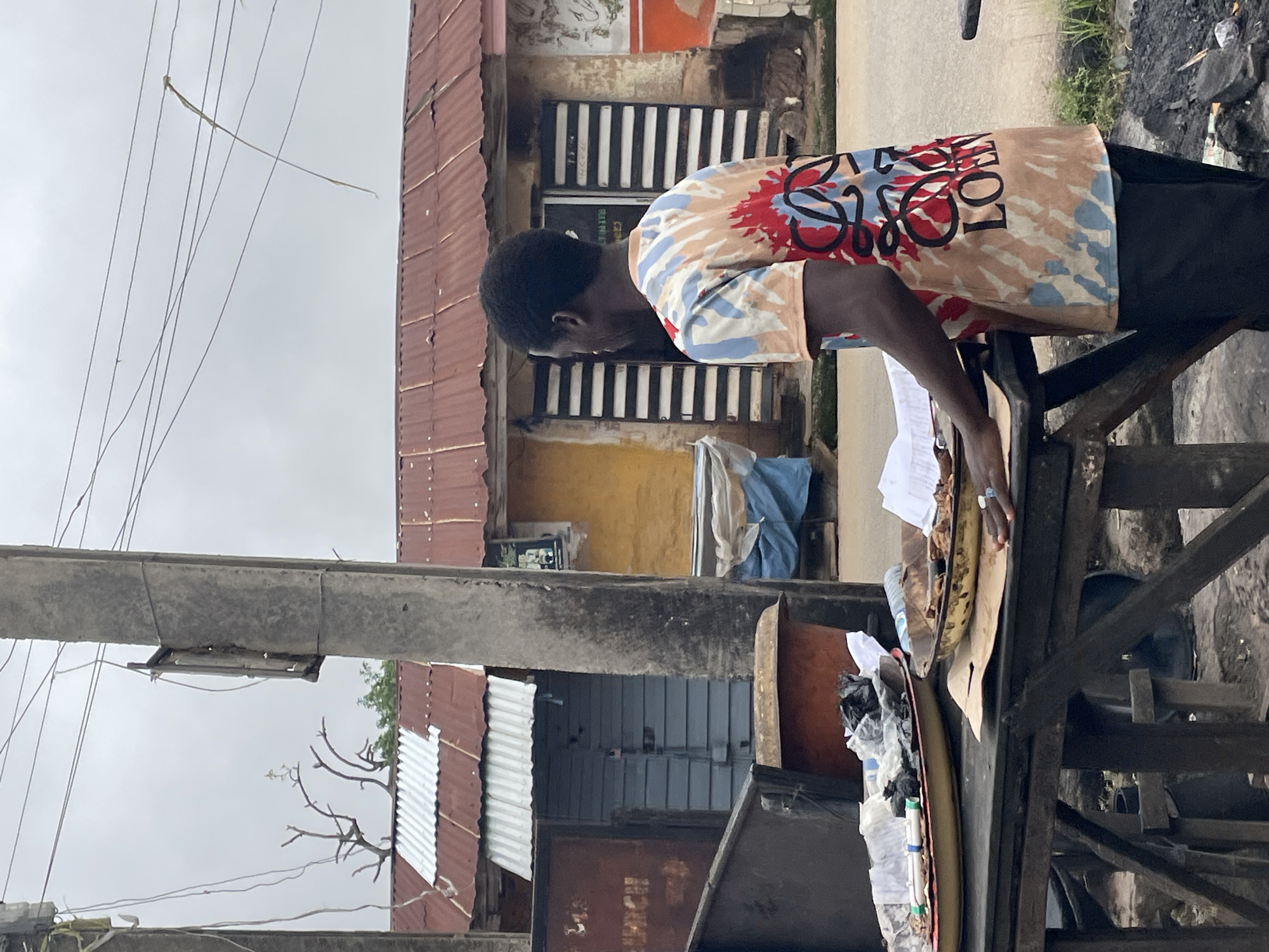 In the city of Jos, Nigeria, a young man with dark skin and short black hair stands behind a wooden table laden with various items. He is wearing a tie-dye t-shirt in shades of beige, blue, red, and black, featuring a large, abstract graphic with the letters "LOEW" prominently displayed. His right hand rests on a metal tray containing what appears to be food items, some of which are covered by paper. A large, shallow metal dish to his left holds crumpled papers, plastic bags, and what seem to be discarded items. The table itself is weathered and appears to be made of rough wooden planks supported by a crude wooden structure.
Behind the man, a building with a corrugated iron roof, a mix of red and silver, stretches across the background. The building has yellow and white painted walls, with dark horizontal slatted windows. A section of the roof is covered by a white corrugated material. To the left of the building, a grey corrugated metal structure is visible. In front of the building, a blue tarpaulin is draped over some objects, and a weathered orange basin sits on the ground.
The scene is framed by two tall concrete utility poles. The pole on the left has wires attached, and a small wooden fixture is mounted on it. The pole on the right also has wires extending from it, with some trailing loosely. The ground in the foreground appears to be dirt and scattered debris, with a pile of dark material, possibly coal or ash, to the right of the table. The sky is overcast and grey.