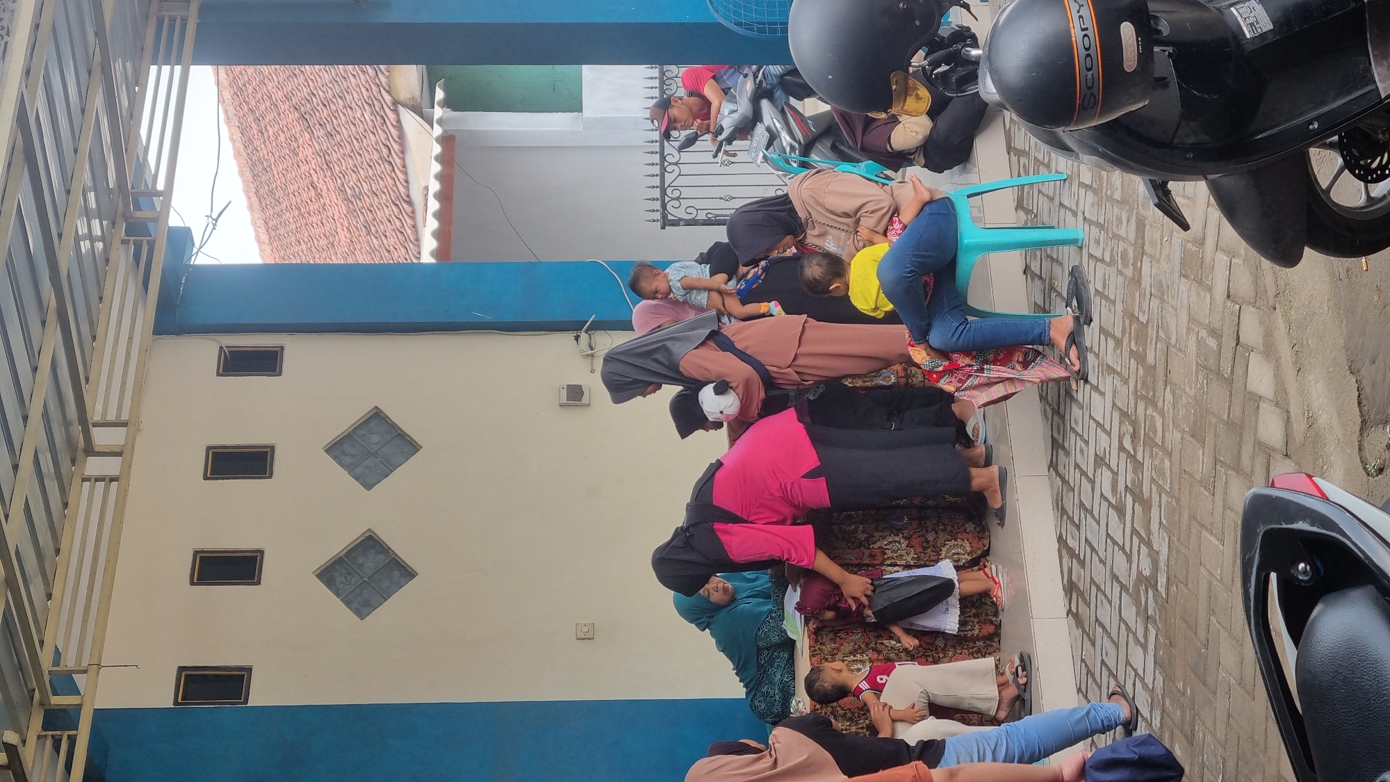 The image depicts an outdoor scene, likely in Diwek, Indonesia, featuring a group of people, predominantly women and children, gathered under a covered walkway. The architecture suggests a public building, possibly a community center or clinic, with a white facade, blue trim, and a metal roof supported by posts. Several windows with dark frames and some diamond-shaped decorative elements are visible on the wall.
In the foreground, two motorcycles are parked, one black with a "Scoopy" logo on the helmet, and another partially visible on the left. The ground is paved with light-colored bricks.
The central focus is the gathering of people. A woman in a bright pink top and dark headscarf is bending over, interacting with a small child in a white dress and red shoes. To her left, a woman in a teal headscarf and dress is seated, seemingly attending to something on a table covered with a patterned cloth. Several other women and children are present. One woman in a light brown headscarf and outfit holds a baby, and another woman in similar attire is seated on a light blue chair with a child in a yellow shirt on her lap. Other children are standing or being held. A man in a red shirt and a cap is seated on a motorcycle in the background, observing the scene. The overall atmosphere appears to be one of casual social interaction or a waiting area.