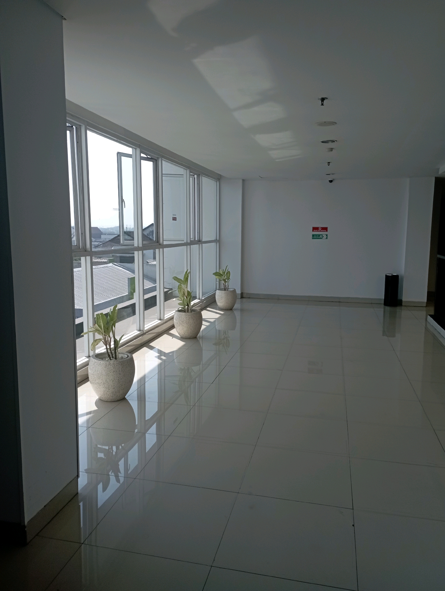The image depicts an interior hallway with large windows on the left side, offering a view of buildings and a clear sky. The hallway features a polished tile floor that reflects the light from the windows and ceiling. Several potted plants are placed along the window wall, adding a touch of greenery to the space. On the right wall, there are two signs, one red and white with text, and a green exit sign pointing to the right. A black trash receptacle is positioned near the wall. The ceiling has a clean, white finish and is fitted with recessed lighting fixtures and what appear to be smoke detectors and sprinkler heads. The overall impression is one of a modern, well-lit, and clean indoor environment. The scene does not contain any people or explicit indications of activity. Based on the information provided and the visual cues, it's plausible this image was taken in a building in the city of Malang, Indonesia, given the architectural style and potentially the vegetation visible through the windows.