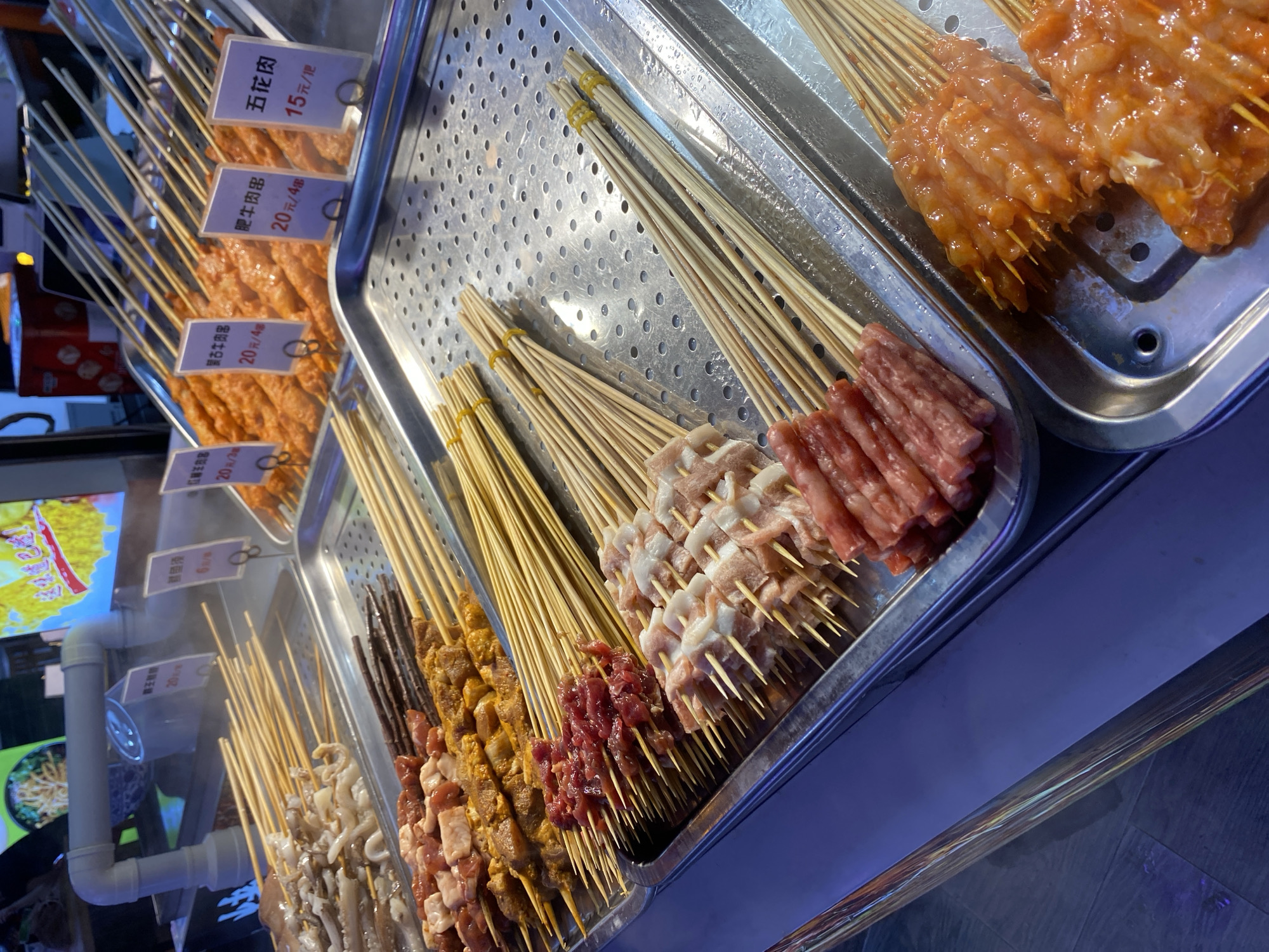 The image shows a close-up, eye-level, indoor shot of a food stall in the Yuzhong District of China, likely a street food market. The stall features numerous skewers of uncooked meat and seafood, displayed in metal trays. The items are prepared for grilling. From front to back, and left to right, the skewers appear to include sliced pork belly, raw beef cubes, possibly chicken or pork pieces marinated in an orange sauce, and what looks like seafood on the far left. Small paper labels are placed in front of some of the skewers, indicating prices and item names in Chinese characters. The lighting is artificial and appears to be from overhead fluorescent lights and possibly some neon signs visible in the background, casting a blue and purple hue on the reflective surfaces. The overall environment is a commercial food vending area, with a focus on raw ingredients ready for immediate cooking.