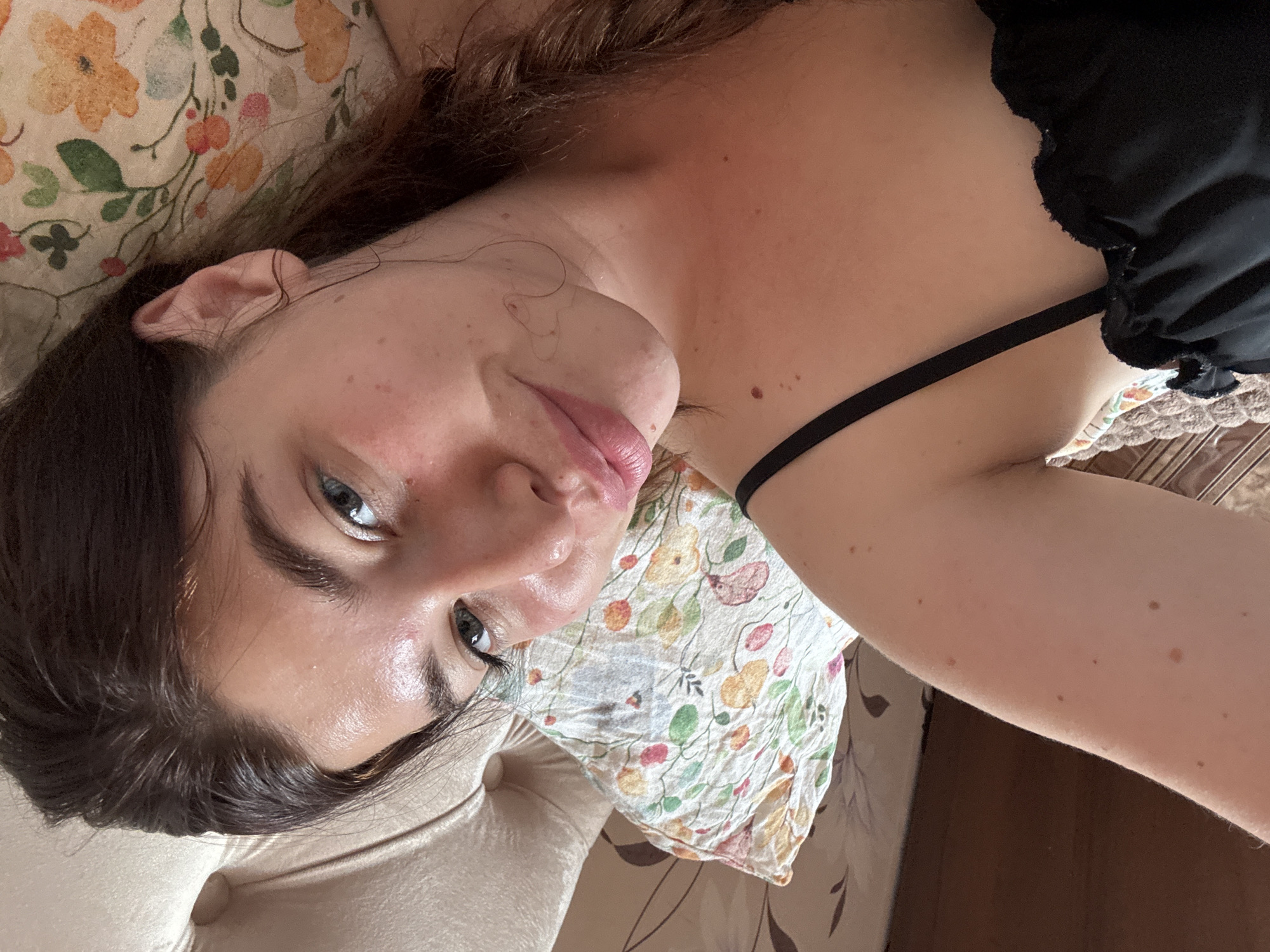 A close-up, overhead shot captures a young woman lying on a bed, with her head resting on a pillow. She has dark hair pulled back into a braid and is looking at the camera with a slight smile. Her skin appears natural and dewy, with a few scattered freckles and moles visible. The pillow beneath her head has a floral pattern in shades of pink, orange, and green. The background of the image shows a hint of a tufted headboard and patterned bedding. The overall impression is one of relaxed intimacy. There are no specific details that would allow for the identification of the location in Klin, Russia.