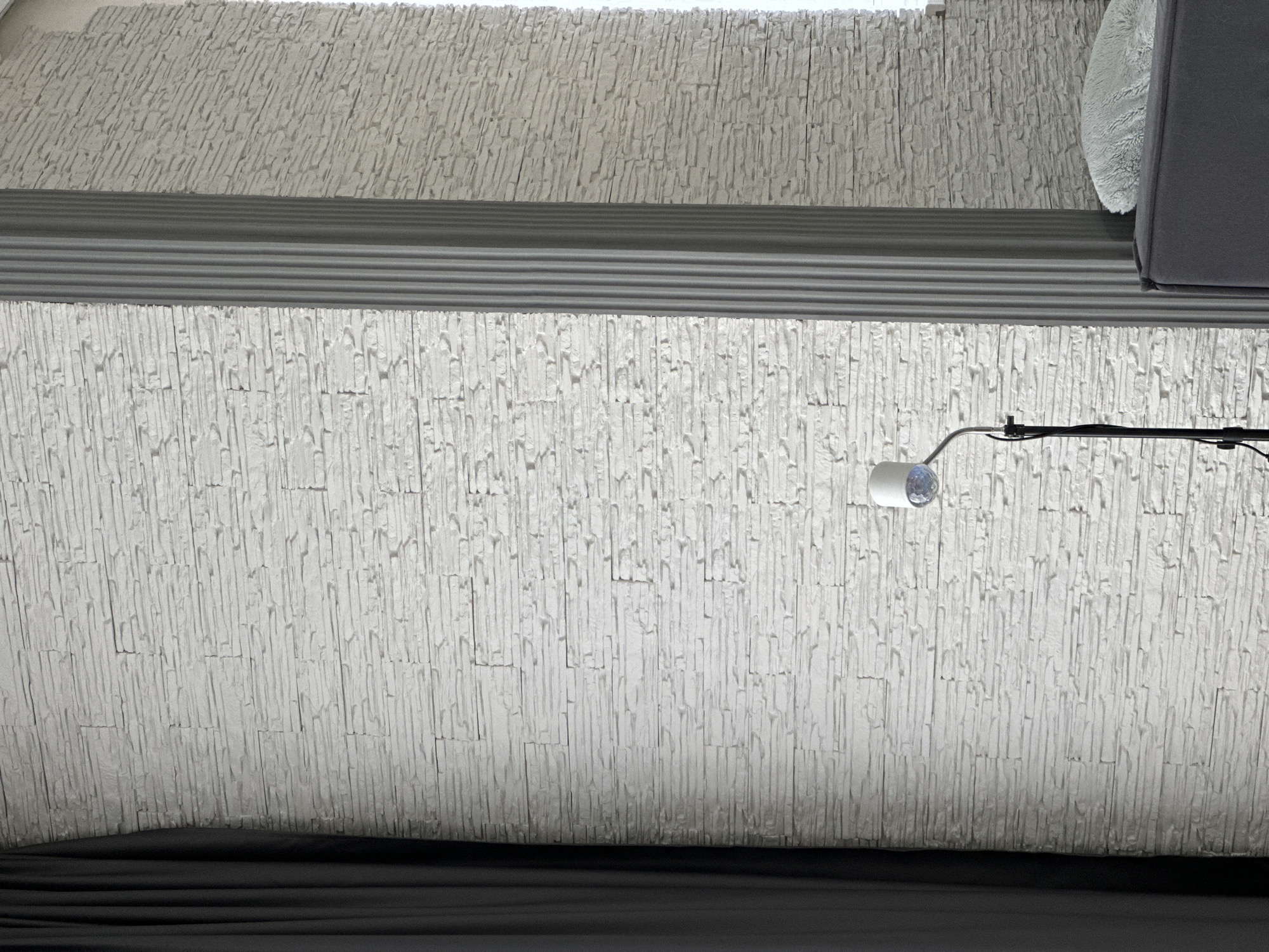 Here is a detailed description of the image:
The image shows a close-up view of a textured white wall, likely an interior space. The wall has a pattern of vertical grooves resembling stacked stones. A gray horizontal band runs across the middle section, creating a division in the pattern.
In the upper right, a portion of a gray object is visible, possibly a piece of furniture or equipment. Near the middle-right, a small, round lamp is attached to the wall with a flexible metal arm. A dark fabric is at the bottom of the image, possibly a curtain or the edge of a piece of furniture.
The location is not specified, so it's only described as "Unknown, Unknown" per instructions.