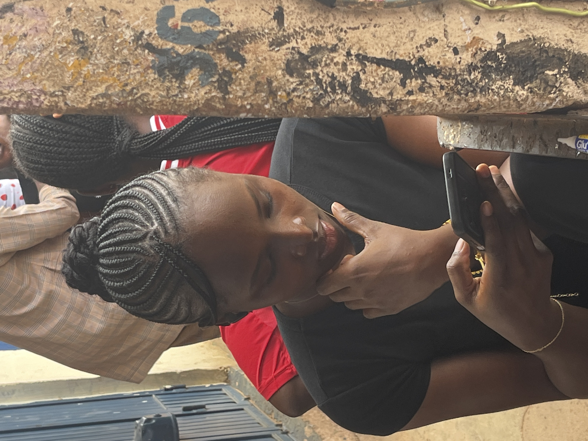 A young Black woman with braided hair pulled into a bun is looking down at her smartphone, holding it in her left hand. Her right hand is touching her chin. She is wearing a black t-shirt and a gold bracelet on her left wrist. In the background, another Black person wearing a red shirt is visible. The scene takes place outdoors next to a textured, graffiti-marked pole. The ground appears to be dirt. The presence of a fence, a blue metal gate, and the general architecture suggest this scene could be located in a residential or commercial area within the city of Jos, Nigeria.