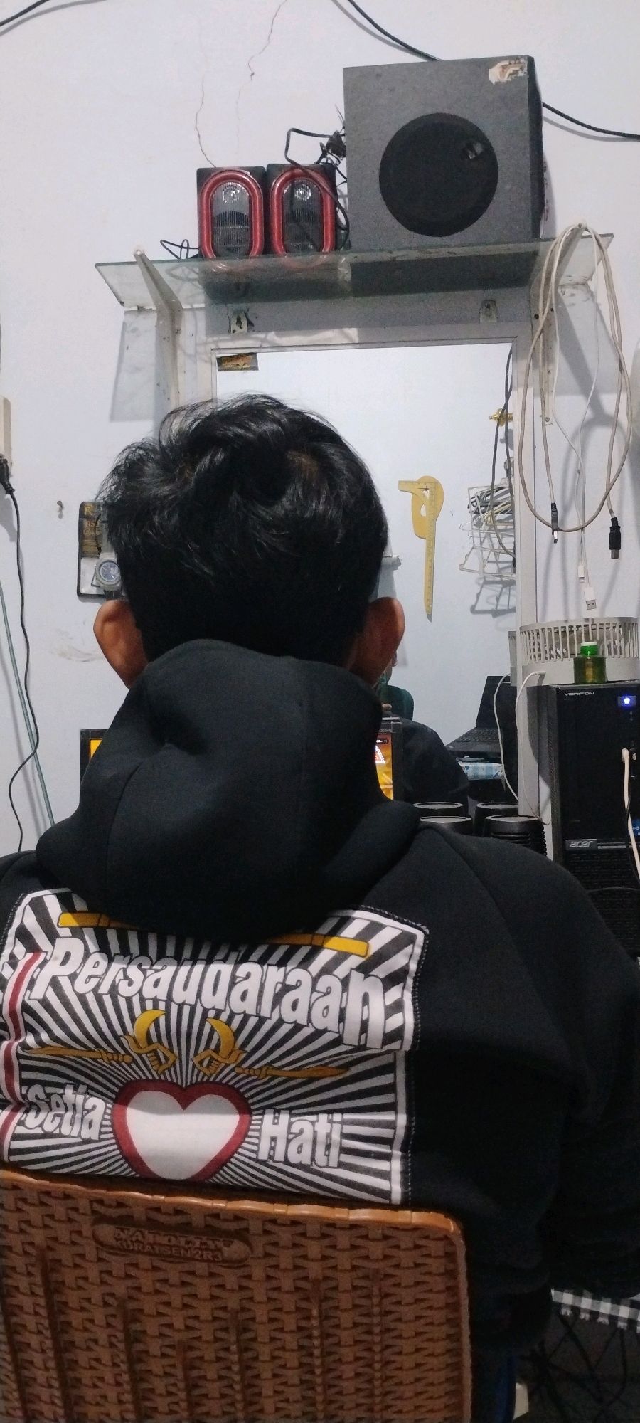 The image shows the back of a person wearing a black hooded sweatshirt with a graphic design on the back. The graphic features text that appears to be "Persaudaraan Setia Hati" which translates to "Brotherhood of the Faithful Heart," a common phrase associated with the Pencak Silat martial art. The person is seated in front of a computer setup. Visible objects include computer monitors, speakers, wires, and a woven basket. The setting appears to be a room within a house, likely in Mumbulsari, Indonesia, as suggested by the presence of the cultural imagery on the sweatshirt.