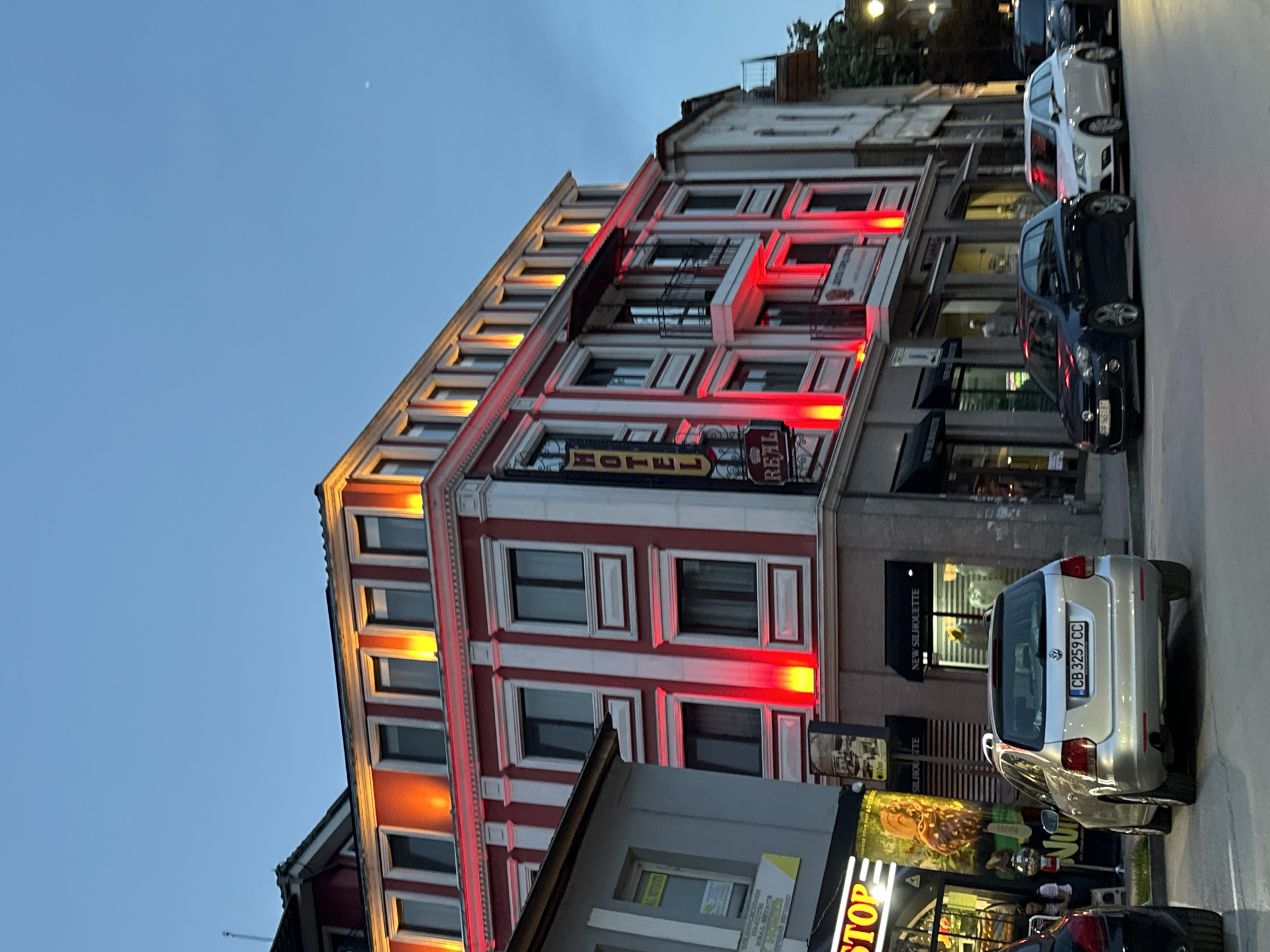 This media file captures an urban street scene at dusk in Veliko Tarnovo, Bulgaria, presented with a 90-degree clockwise rotation. The primary subject is a multi-story building, prominently a "HOTEL REAL," featuring a distinctive red and white facade. The building is illuminated by vibrant red and warm orange strip lights that accentuate its windows and architectural lines, creating an eye-catching glow. A yellow sign clearly spells "HOTEL," while a decorative red sign below it reads "REAL."
Along the street, several cars are parked. A silver Volkswagen hatchback is clearly visible, bearing a Bulgarian license plate "CB 3259 CC," which aligns with the given location context. Further down the street, a dark-colored car and a white car are also parked. Adjacent buildings with storefronts line the street, and one black sign reads "NEW SILHOUETTE."
The sky, visible in the upper portion of the rotated image, is a clear, deep twilight blue, with a faint, small moon discernable. Other visible text includes "STOP" on a shop sign at the bottom, and partially visible words like "REAL ESTATE" on another sign. The overall scene depicts a lively evening atmosphere as artificial lights begin to dominate the fading daylight.