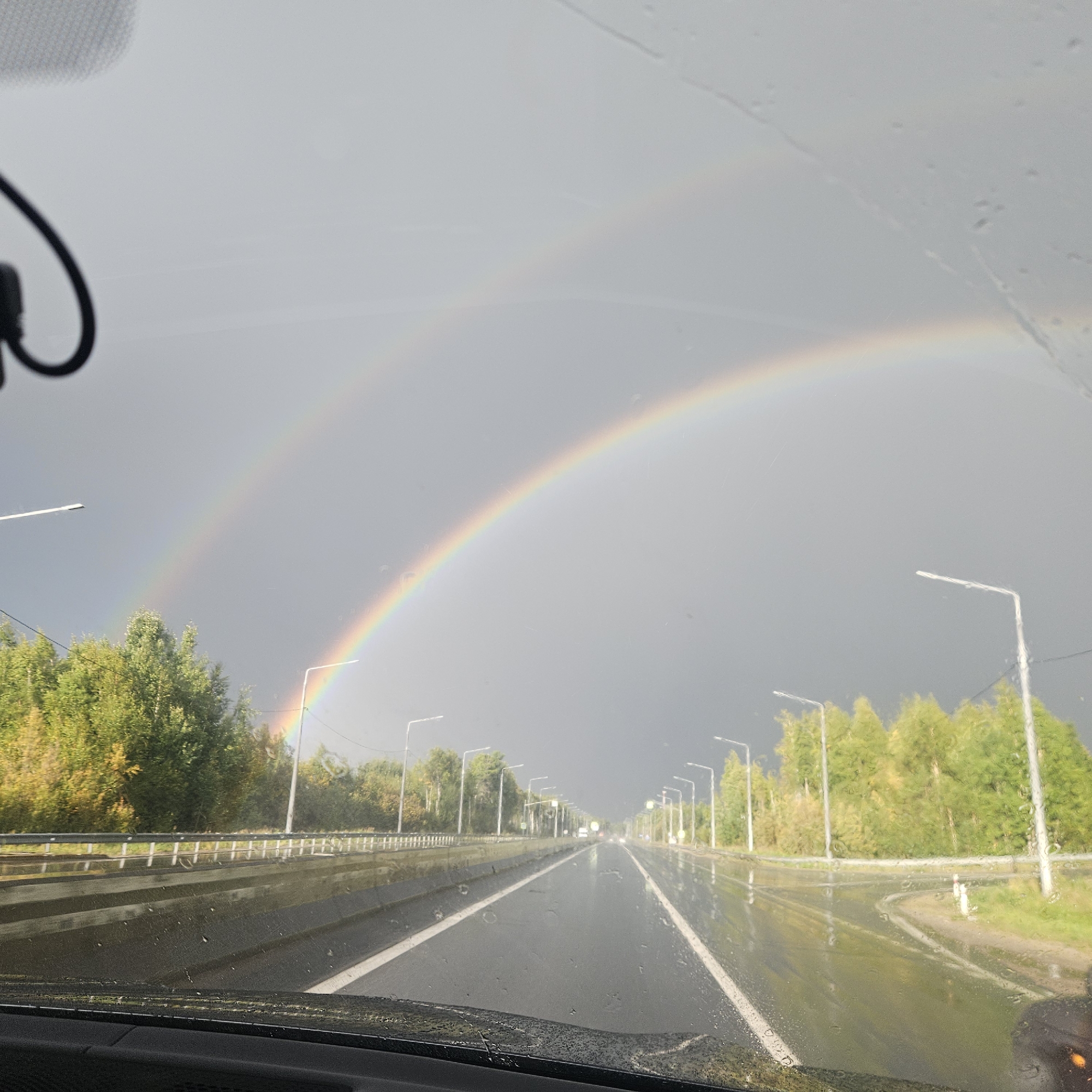 An overcast sky looms over a wet, multi-lane road in Megion, Russia, with a double rainbow arching across the horizon. The road, possibly a highway, is lined with trees and streetlights. The scene appears to be captured from within a vehicle, indicated by the dashboard and windshield in the foreground, which is speckled with raindrops. The overall atmosphere is somber, due to the weather, but illuminated by the vibrant colors of the double rainbow.