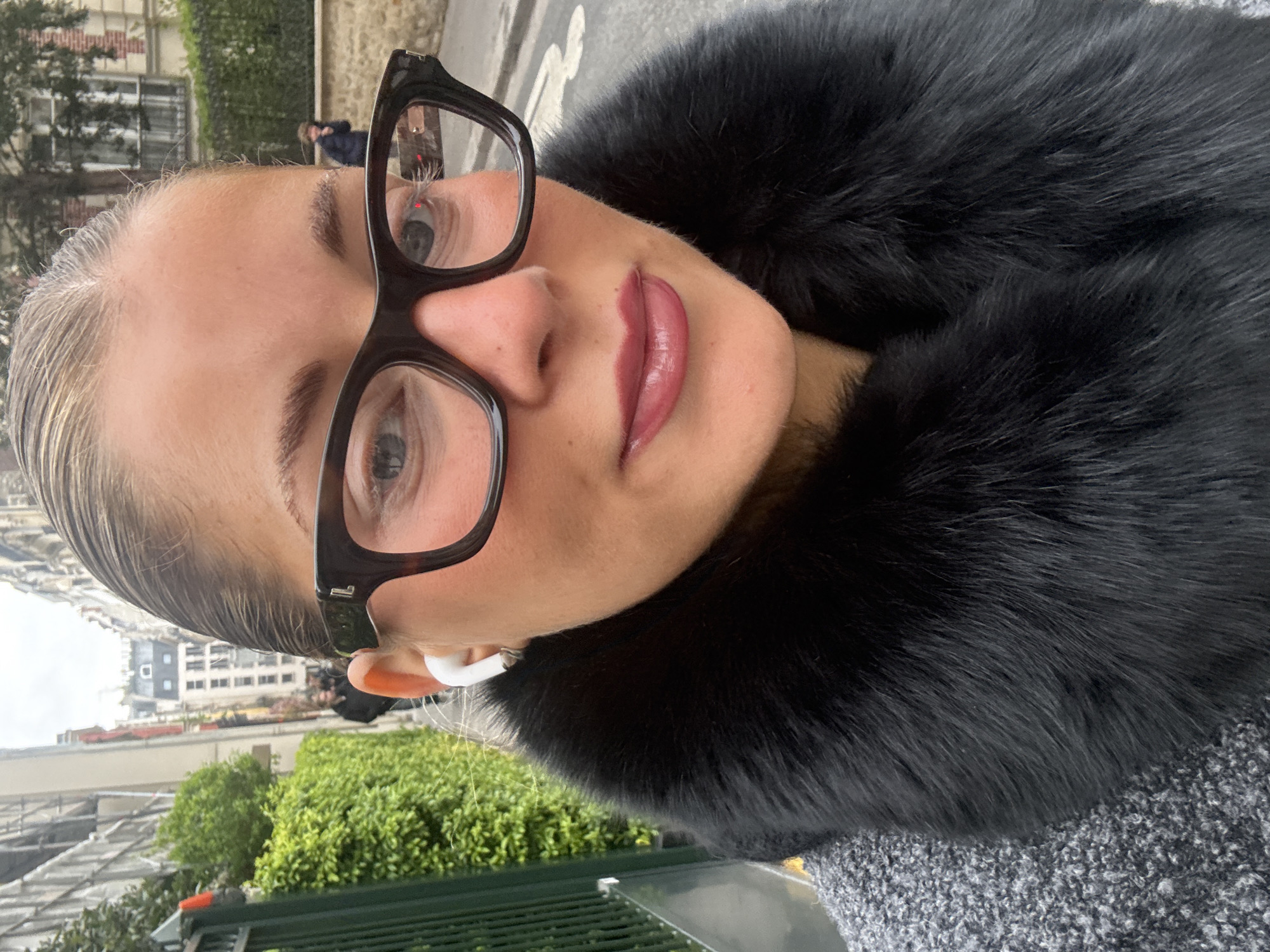 A selfie depicts a woman with light skin, wearing glasses and a dark fur collar. She has slicked-back blonde hair, full lips with a glossy finish, and an earbud in her right ear. The background suggests an urban environment, with buildings typical of Paris, France, indicating a possible location within the city. Trees and a green fence are visible, contributing to the urban landscape. A person is seen walking in the background.