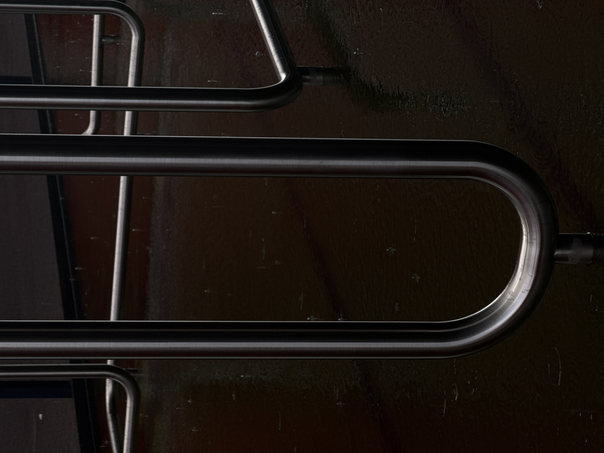 The image shows a close-up of metal railings or barriers against a dark, glossy surface. The railings are made of smooth, rounded metal tubes, possibly stainless steel. The surface behind the railings is dark and reflective, appearing like a varnished or wet surface. The scene is dimly lit, with highlights on the metal indicating a light source. The overall image is abstract and focuses on the textures and forms of the railings and the reflective surface. The location is likely Esch-sur-Alzette, Luxembourg, based on the prompt, but there is no explicit information to confirm this in the image itself.
