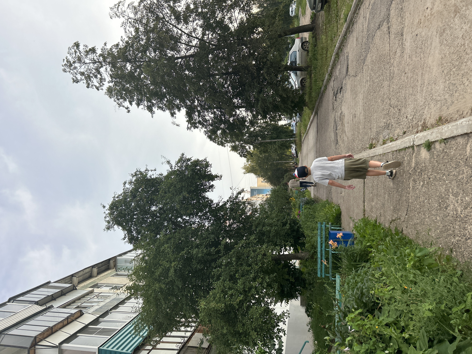 A boy wearing a white t-shirt and green shorts walks down a paved sidewalk in Chaikovsky, Russia. He is wearing a blue baseball cap. He is walking away from the camera, towards a line of trees and parked cars. To the left of the sidewalk is a grassy area with benches and flowering plants. Beyond the greenery is a multi-story apartment building. The sky is overcast.