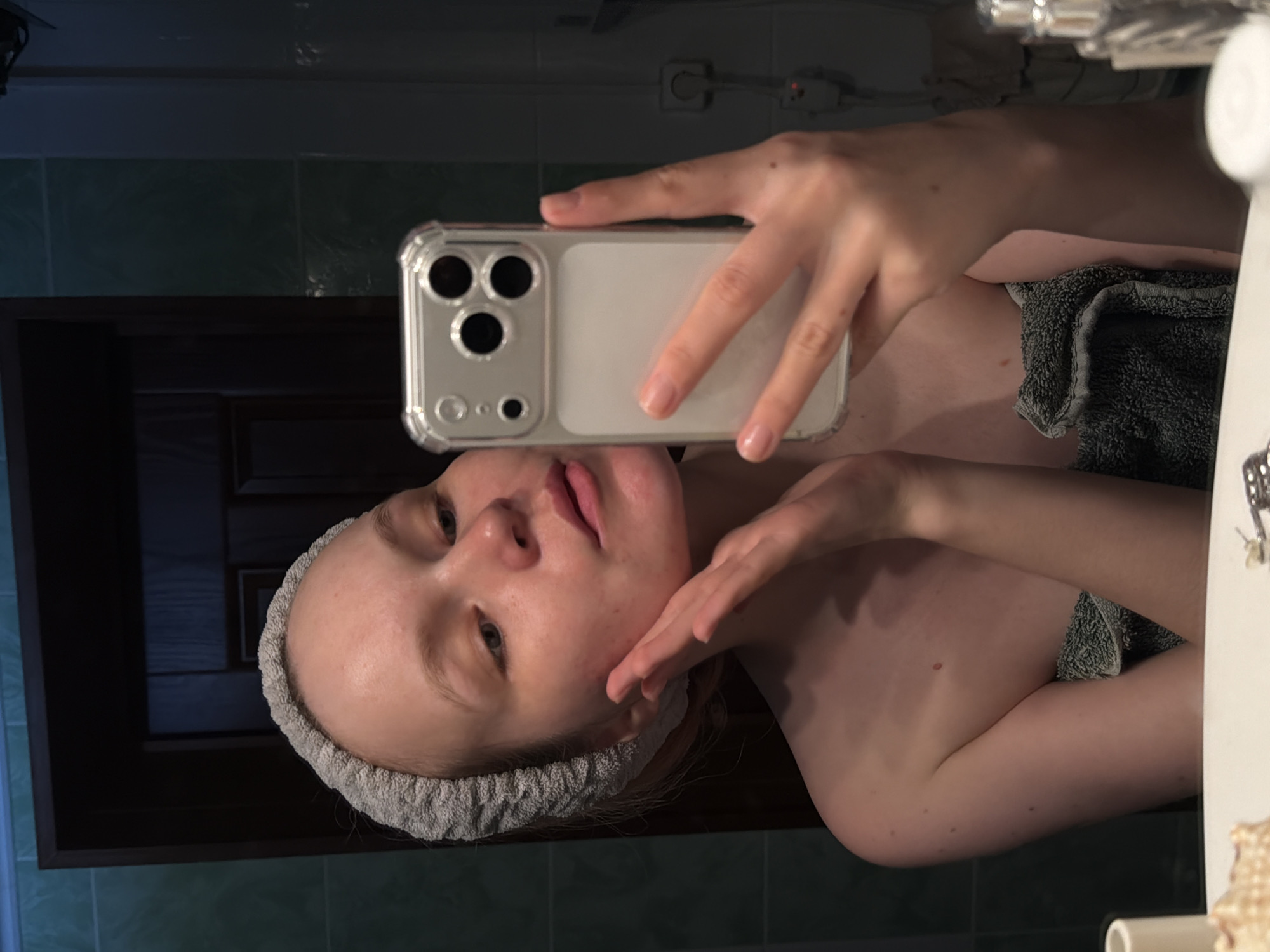 A person with a gray headband is taking a selfie in a bathroom. They are holding a smartphone with a clear phone case. A dark gray towel is draped over their shoulder. The bathroom features dark green wall tiles and a dark wood-colored cabinet door. Кадряково, Russia is not discernible from the image.