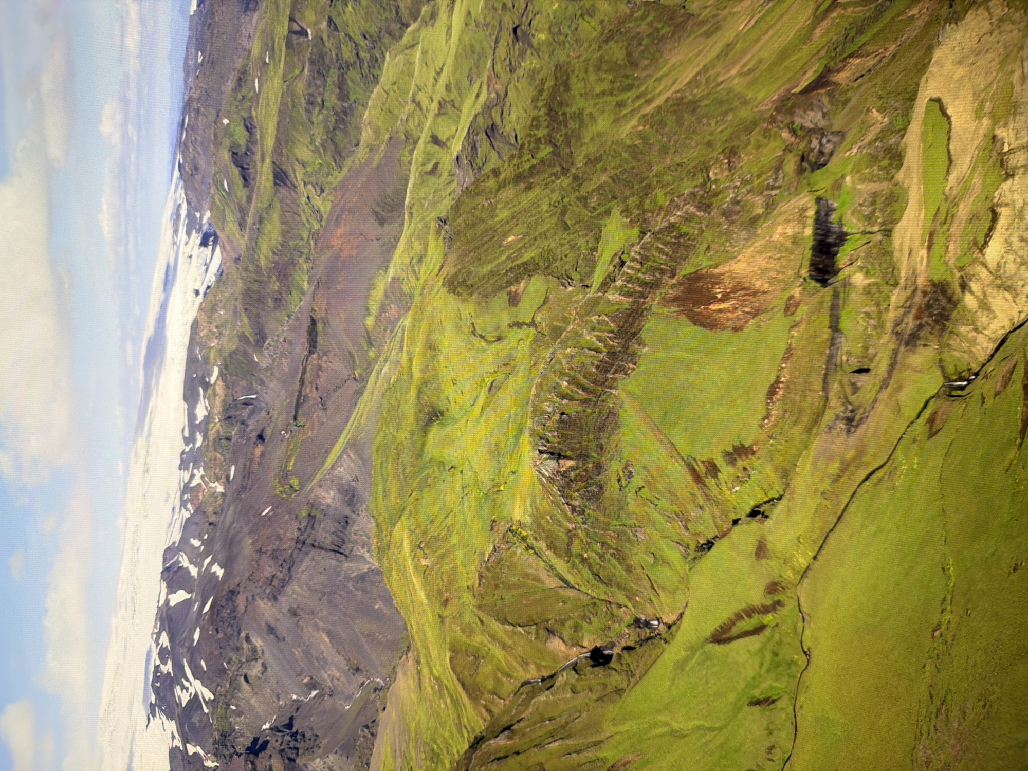 This is an aerial photograph of a dramatic, mountainous landscape. The foreground is dominated by rolling, vibrant green hills and slopes, etched with dark ravines and winding streams or gullies. Some of these ravines appear to have small waterfalls. The terrain is uneven, with sharp inclines and textured surfaces.
In the middle ground, the green hills transition into darker, more rugged, and barren mountain slopes, marked by patches of grey and black rock. These slopes are less vegetated and appear steeper.
In the background, a vast, snow-capped mountain range stretches across the horizon. The snow cover is extensive, suggesting a high altitude or a cold climate. The sky above is a light blue with scattered white clouds, some of which are elongated and wispy, others more puffy.
There are no people, buildings, or any man-made structures visible in this image. The scene depicts a natural, wild environment, likely a remote mountainous region. The image is not of Kyiv, Ukraine.