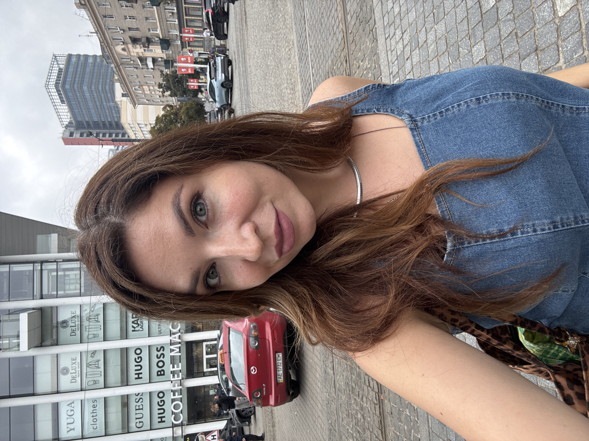 A woman with long, brown hair and light blue eyes poses for a selfie outdoors on a cloudy day. She is wearing a denim dress and a silver necklace. In the background, a red taxi with a license plate that reads "AE 1732 EM" is visible, parked on a cobblestone street. Behind the taxi, a modern building with large glass windows displays various brand names including "YUGA," "DELUXE MEN'S SHOP," "GUESS," "HUGO," "BOSS," and "COFFEE MAC." Further in the background, other buildings and cars, including a blue electric vehicle, are present. The scene appears to be in a city center, likely in Dnipro, Ukraine, given the presence of Ukrainian license plates and architecture consistent with Eastern European cities.