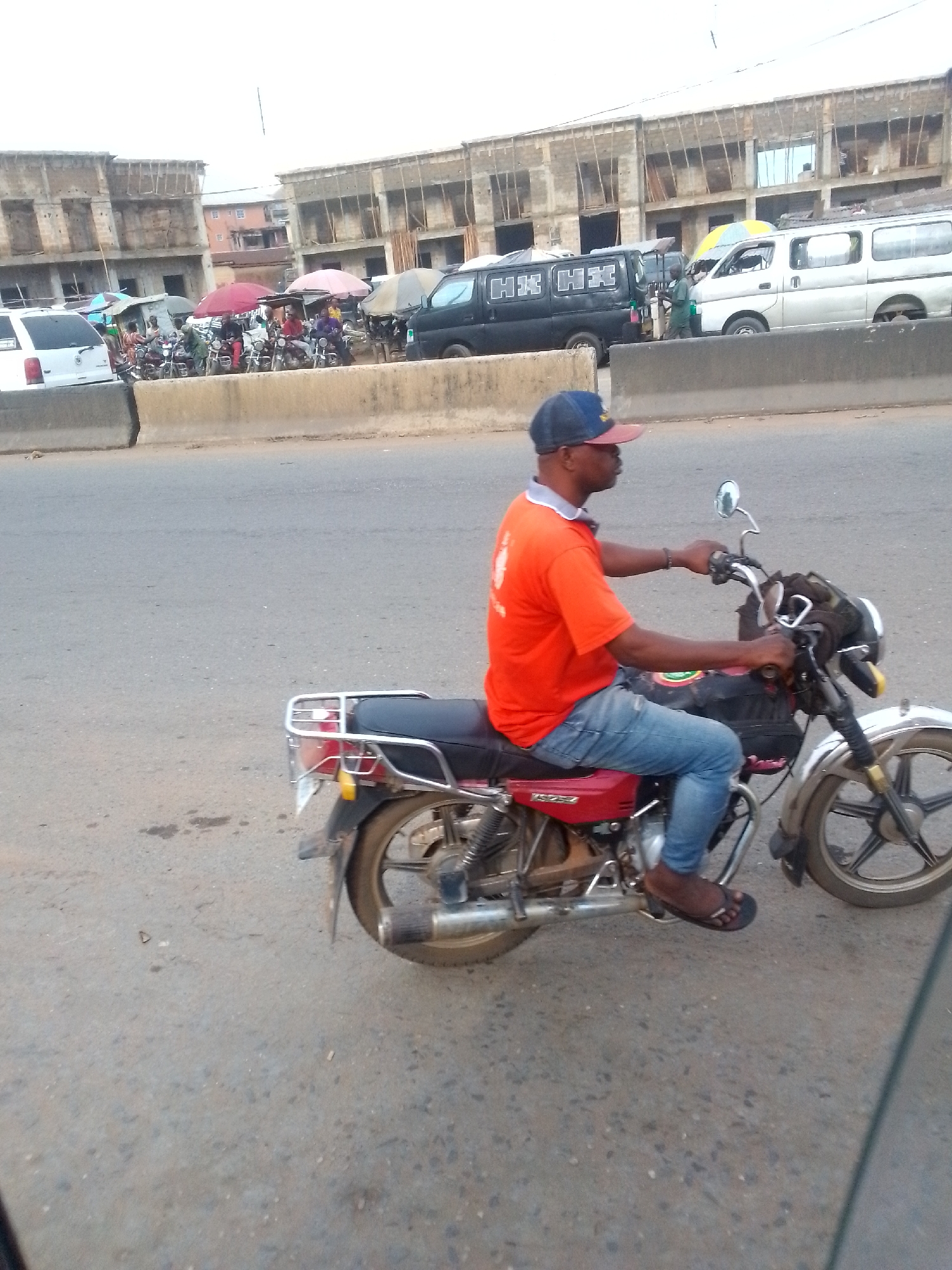 A man rides a red motorcycle on a paved road in Ihiala, Nigeria. He is wearing an orange t-shirt, blue jeans, and a blue baseball cap with a red bill, and is shod in flip-flops. The road is gray asphalt. In the background, across a concrete barrier, are buildings under construction, vehicles (vans, cars, motorcycles) and people near umbrella-shaded stands. The sky is overcast.