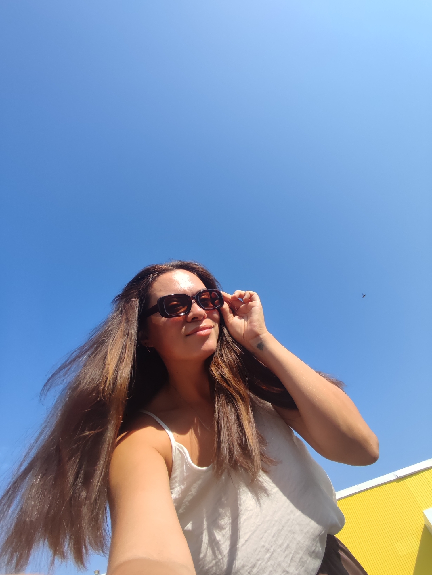 In a low-angle shot against a bright blue sky, a woman with long, brown hair is captured outdoors, possibly in Chaikovsky, Russia, given the context. She is wearing dark, rectangular sunglasses and a white tank top. Her right hand is raised, adjusting the sunglasses, and a small tattoo is visible on her inner wrist. A tiny dark speck, possibly a bird, is in the upper right quadrant of the sky. In the lower right corner, a section of a building with vibrant yellow siding is visible, suggesting an urban or park setting. The overall lighting indicates a sunny day.