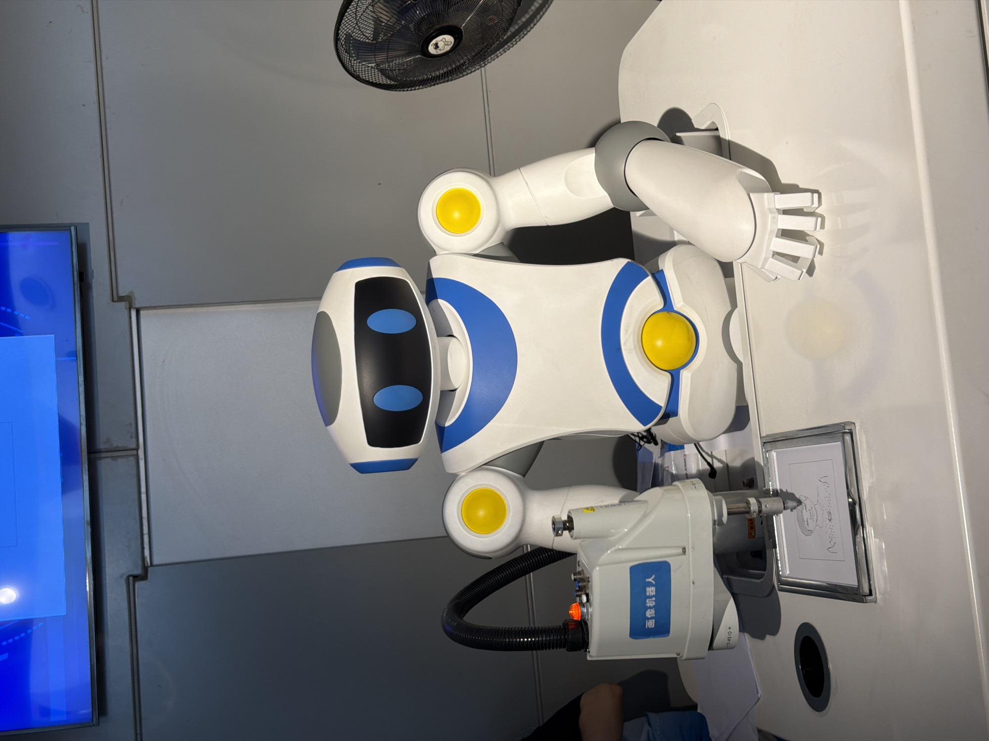 This image depicts a white and blue robot with a gray head and yellow accents, positioned at a white table. The robot is actively drawing a portrait on a framed piece of paper with a pen attached to its arm. To the left of the robot, there is a white machine with Chinese characters, labeled "画像机器人" (Portrait Robot), which appears to be controlling the drawing process. The robot's other arm is extended, with its hand resting on the table. In the background, a large screen displays a blue image, and to the right, a black oscillating fan is visible. The scene is likely within an exhibition or demonstration setting.