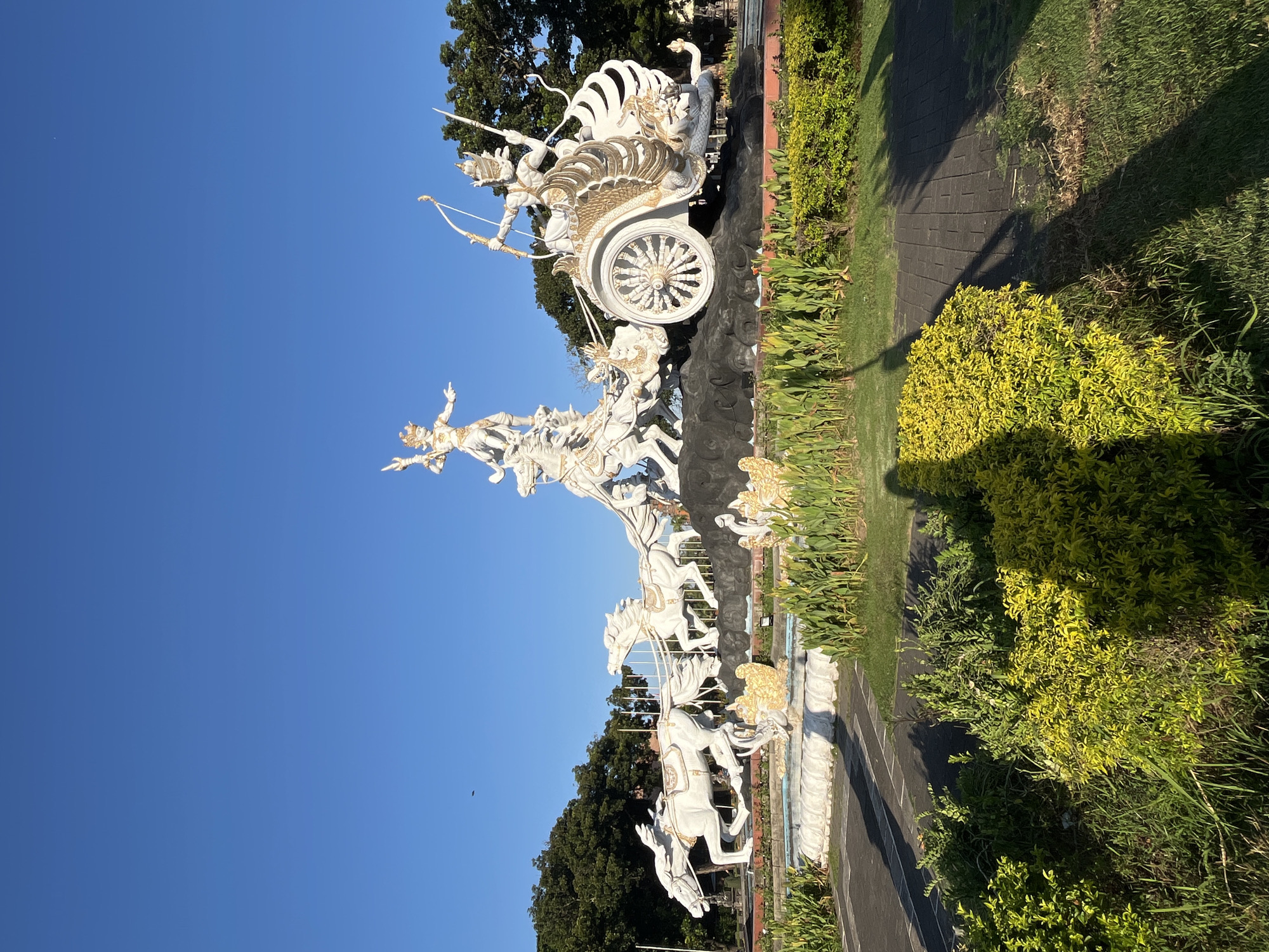 This is a photograph of the Puputan Monument in Kuta, Bali, Indonesia. The monument depicts a dramatic scene of a chariot pulled by horses, with figures in traditional attire. The central figure in the chariot is armed with a bow and arrow, and another figure stands tall on a horse in front of the chariot. The monument is rendered in white with gold accents, creating a striking contrast against the bright blue sky. It is surrounded by lush green foliage and a fountain, with a paved walkway in the foreground. The sunlight casts long shadows across the scene, indicating it is late afternoon. The monument commemorates the mass suicide or "puputan" that occurred in Bali during the Dutch colonial era.