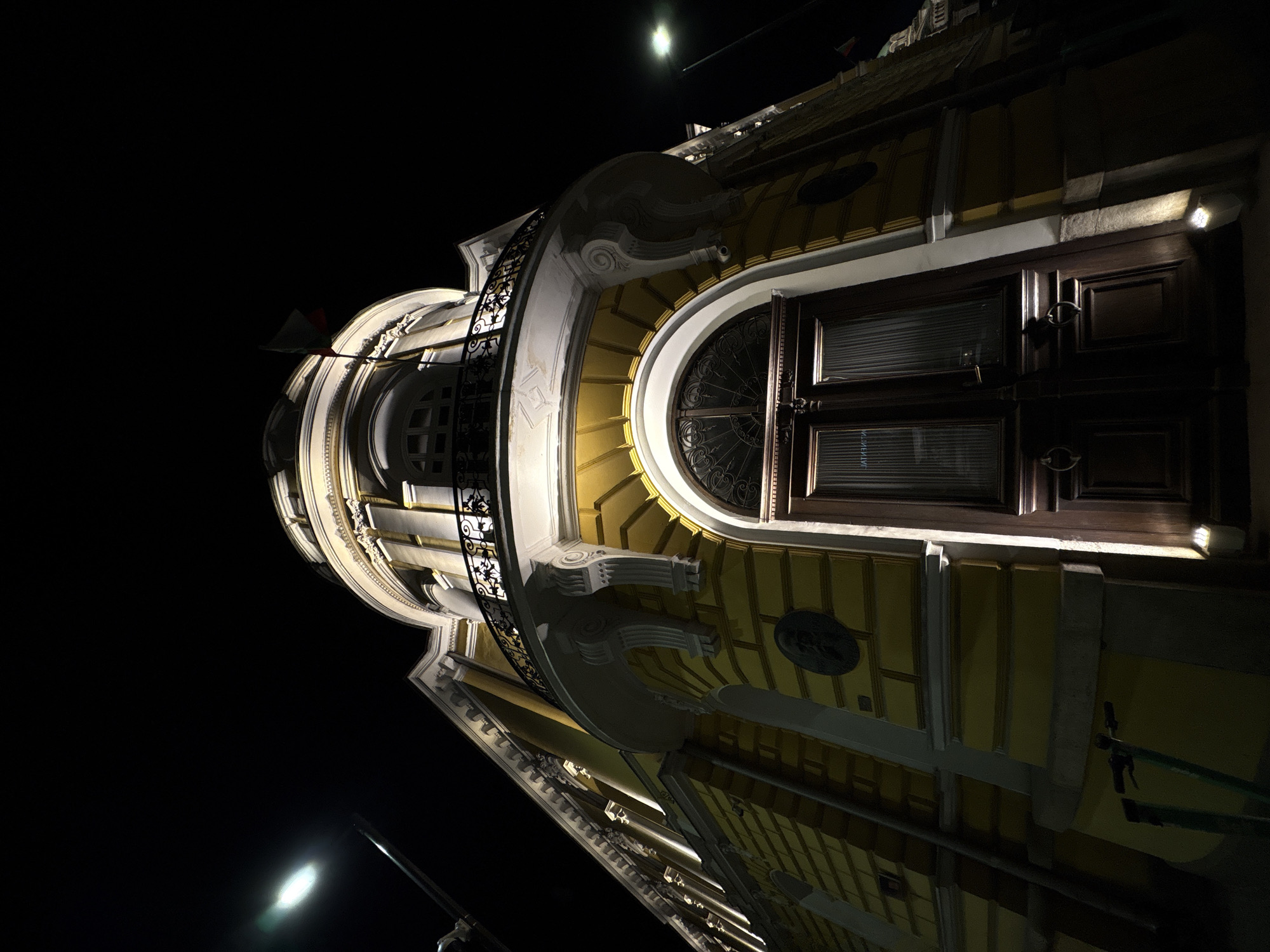 This image presents a dramatic low-angle view of an ornate building facade in Sofia, Bulgaria, captured at night. The dark, featureless sky emphasizes the illuminated architectural details of the structure.
The building displays a blend of classical and possibly Baroque influences, characterized by a light yellow or cream color scheme on its main surfaces, contrasted with bright white decorative elements. Prominent features include a large, dark wooden arched double door or window frame with glass panels and internal blinds on the right, and another smaller, circular window to the left. The facade is richly decorated with intricate moldings, cornices, scroll-like carvings, and ornate support brackets. A decorative wrought-iron balcony railing is visible on the left side of the building.
The structure is brightly lit by artificial lights, creating strong highlights on the white architectural details and casting deep shadows that accentuate its three-dimensional form. Two bright streetlights are visible as luminous orbs against the black sky – one at the top-center and another towards the bottom-left. A small flag, possibly displaying red and green colors, is partially visible near the top-left edge of the building. Above the main arched door/window, a small, modern-looking white light fixture or sensor is attached to the wall.
Faint, reversed text, difficult to decipher, is visible on the glass panels of the main doors, likely a reflection or internal signage. The scene evokes a sense of historical grandeur and architectural beauty, highlighted by the nighttime illumination.