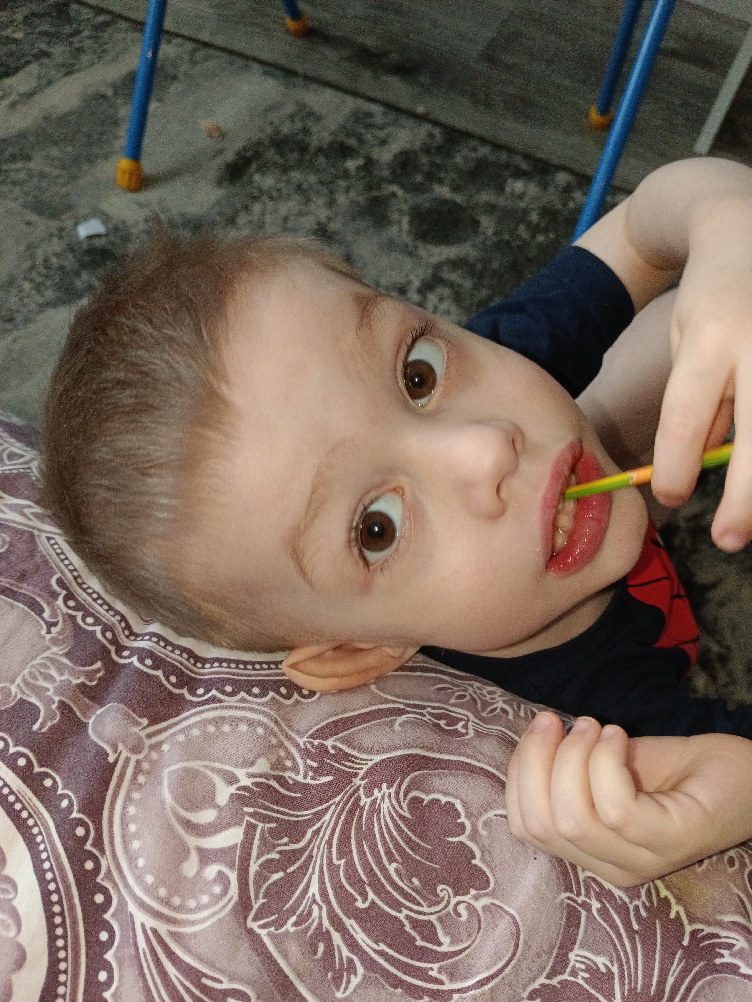 The scene is located in Chaikovsky, Russia. A young child with light brown hair, fair skin, and wide brown eyes looks directly at the camera. The child is lying on their left side, resting on a patterned fabric with white and light purple paisley-like designs on a darker purplish-brown background. They are wearing a dark blue shirt, which appears to have a red detail visible on the right shoulder area. The child holds a multi-colored object, appearing to be a toothbrush with green, yellow, and orange sections, in their right hand, with one end inserted into their slightly open mouth. Their lower lip is visibly moisturized or slightly chapped. The background features a dark, textured floor, possibly linoleum or tile, and partially visible blue metal furniture legs with yellow caps. A small white object is present on the floor near the blue legs.