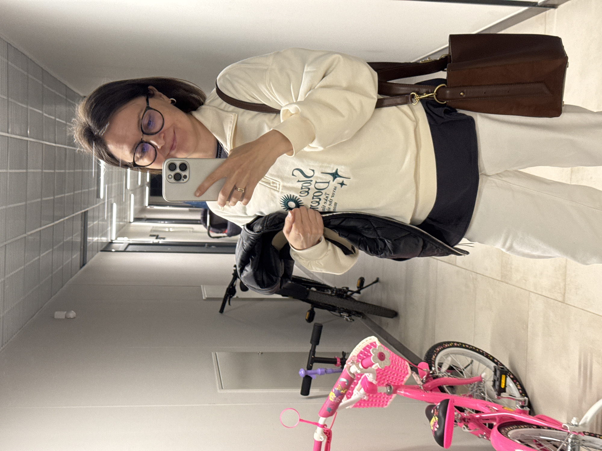 A woman is captured in a mirror selfie within an indoor hallway, likely in a residential building. She has shoulder-length dark hair, wears glasses, and a light-colored (cream/off-white) long-sleeved top with a dark blue collar/undershirt, and white pants. On her left ring finger, a ring is visible. She holds a light-colored smartphone (likely an iPhone) up to take the photo, displaying a slight, pleasant smile.
Her top features mirrored text that reads "Ѕтᴀя Доґя / Star Dog" and a sun-like graphic with rays. Below this, smaller, mirrored text is partially visible, appearing to include words like "IT'S THE ONLY THING THAT MATTERS." She also carries a dark brown leather shoulder bag with gold-colored hardware slung over her shoulder and holds a black padded jacket over her arm.
The setting is a plain, well-lit hallway with light-colored walls and floor. The left wall has a textured or paneled appearance with horizontal lines. Linear light fixtures are reflected in the surface where the photo is taken. In the background, an adult black bicycle is partially visible. More prominently in the foreground and mid-ground are a bright pink child's bicycle with a front basket and training wheels, and parts of a pink scooter. The arrangement of the bicycles suggests a shared storage area or common space within the building. Based on the provided context, this scene is set in Izhevsk, Russia.