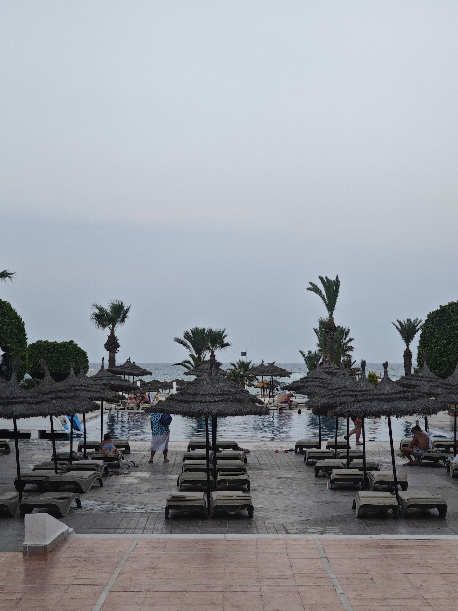 The image depicts a resort swimming pool area in Hiboun, Tunisia, under an overcast sky. In the foreground, rows of empty lounge chairs are neatly arranged on a tiled patio. Further back, a large swimming pool is visible, with several people relaxing on lounge chairs and swimming. Palm trees and thatched umbrellas dot the landscape, providing shade. The sea is visible in the distance. A person in a blue and white patterned garment stands near a child sitting on a lounge chair. Another person, a man, is seated on a lounge chair by the pool. The scene suggests a tranquil resort atmosphere.