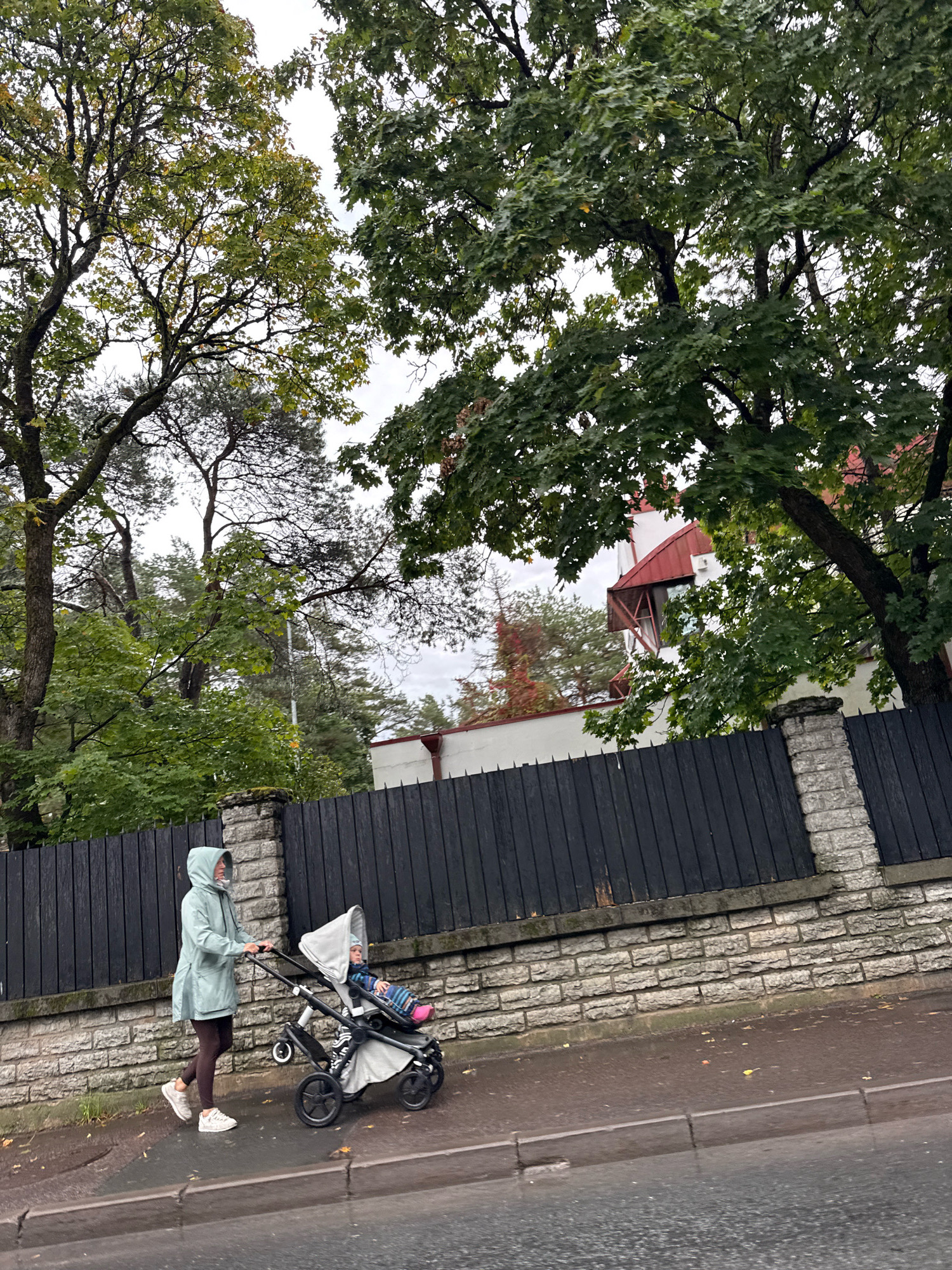 A woman wearing a mint green hooded jacket and dark leggings pushes a baby stroller on a wet sidewalk in Tallinn, Estonia. The stroller is gray and appears to contain a baby wearing a colorful outfit. A dark wooden fence with stone pillars runs along the sidewalk. Tall trees with green leaves are visible behind the fence, and a white building with a red roof is partially obscured by the trees. The sky is overcast, suggesting a cloudy day. The scene depicts a person with a child on a walk in an urban environment with ample greenery.