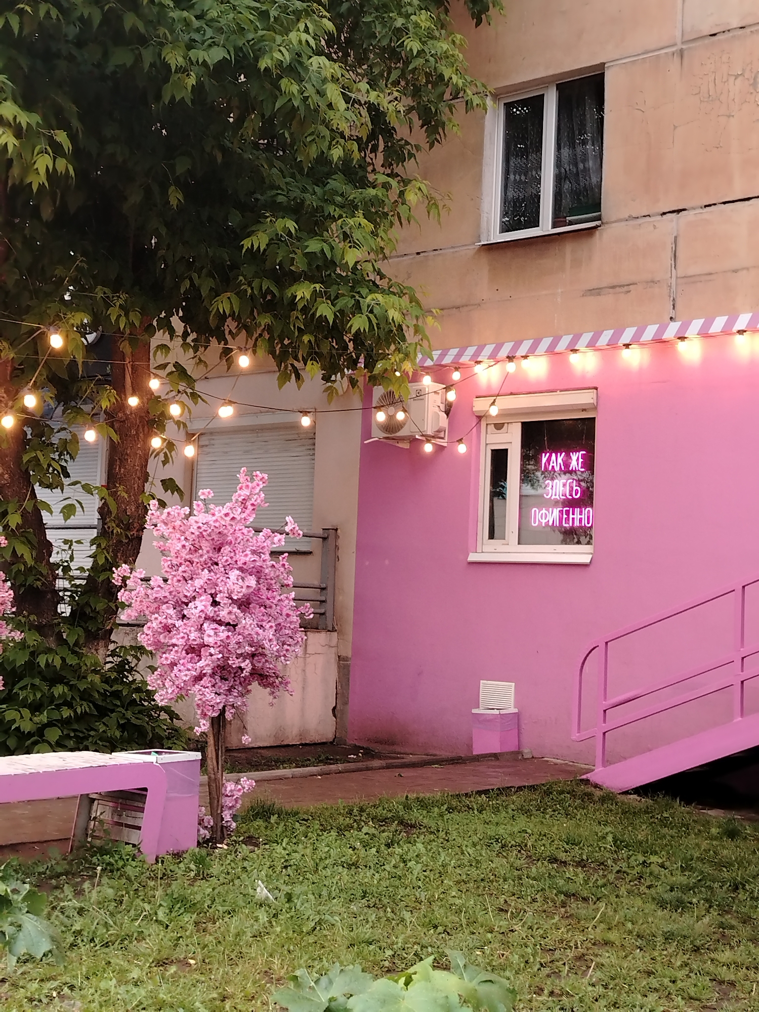 The image depicts an inviting and vibrantly decorated outdoor area, likely an entrance or patio space, next to a building in Izhevsk, Russia. The lower part of the building is painted a striking bright pink, contrasting with the upper floor, which appears to be an older, beige-colored concrete structure with a typical window.
String lights with warm, glowing bulbs are draped across the top of the pink facade, adding to the evening or nighttime ambiance. A white-framed window on the pink wall features a prominent pink neon sign inside, displaying the Russian text: "КАК ЖЕ ЗДЕСЬ ОФИГЕННО." This translates to "It's so awesome here" or "How amazing it is here," conveying a sense of excitement and approval. An air conditioning unit is mounted above the window, and a set of pink stairs or a ramp leads down from the building on the right.
To the left, a large tree with dense green foliage frames the scene. Beneath it, a smaller, artificial-looking cherry blossom tree with an abundance of vivid pink flowers enhances the whimsical theme. A pink bench is positioned on the left side, complementing the overall color scheme. The foreground consists of a grassy patch with some scattered dirt. The lighting, with the illuminated string lights and neon sign, suggests it is either dusk or night, creating a cheerful and eye-catching atmosphere.
