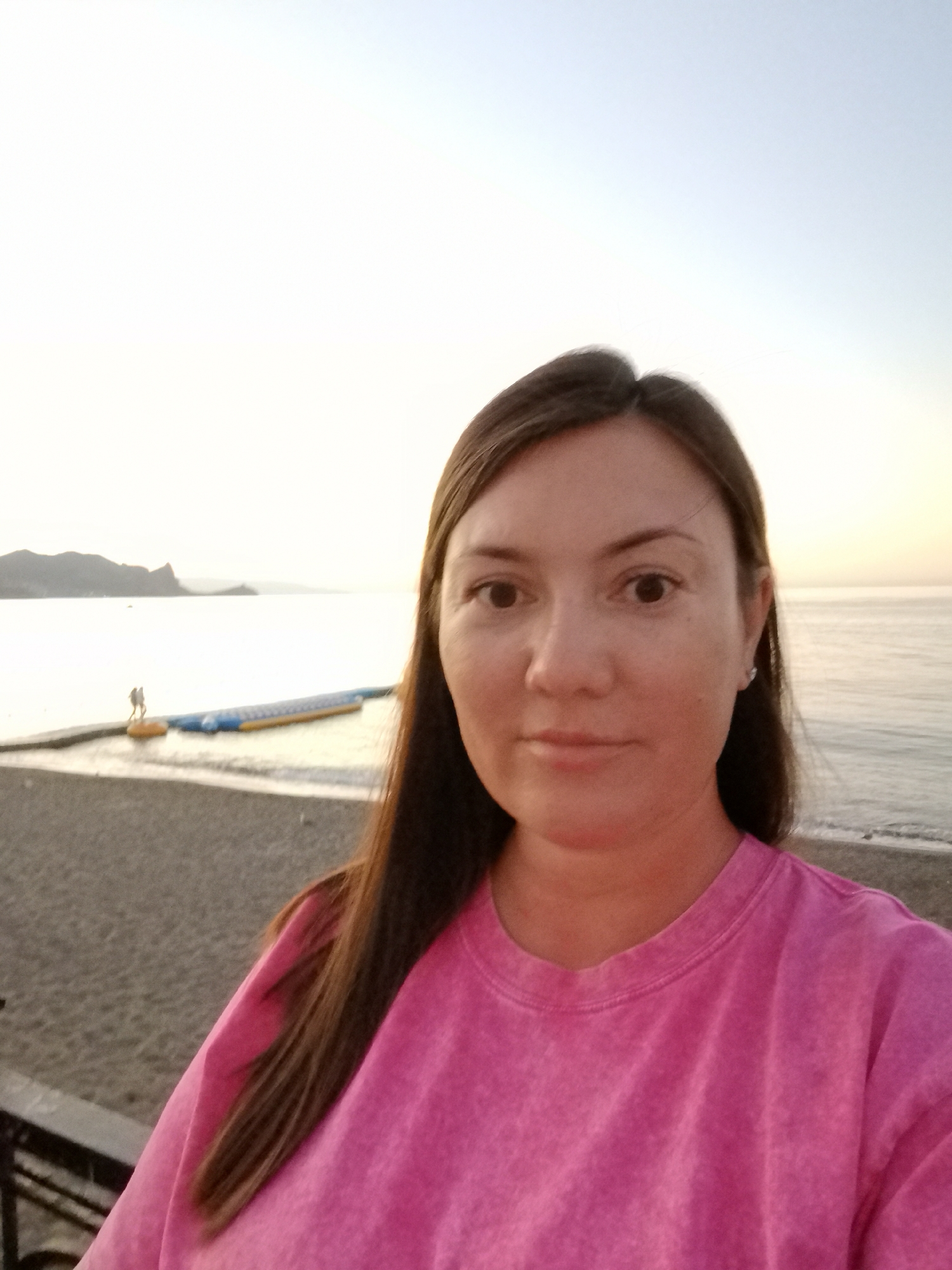 A selfie of a woman in a pink shirt is taken on the beach in Sudak, Ukraine. The woman is looking at the camera with a neutral expression. Behind her, there is a beach with pebbles and a body of water, possibly the Black Sea. In the distance, there are mountains and a floating water attraction. The sky is light and clear, suggesting it is either early morning or late afternoon.
