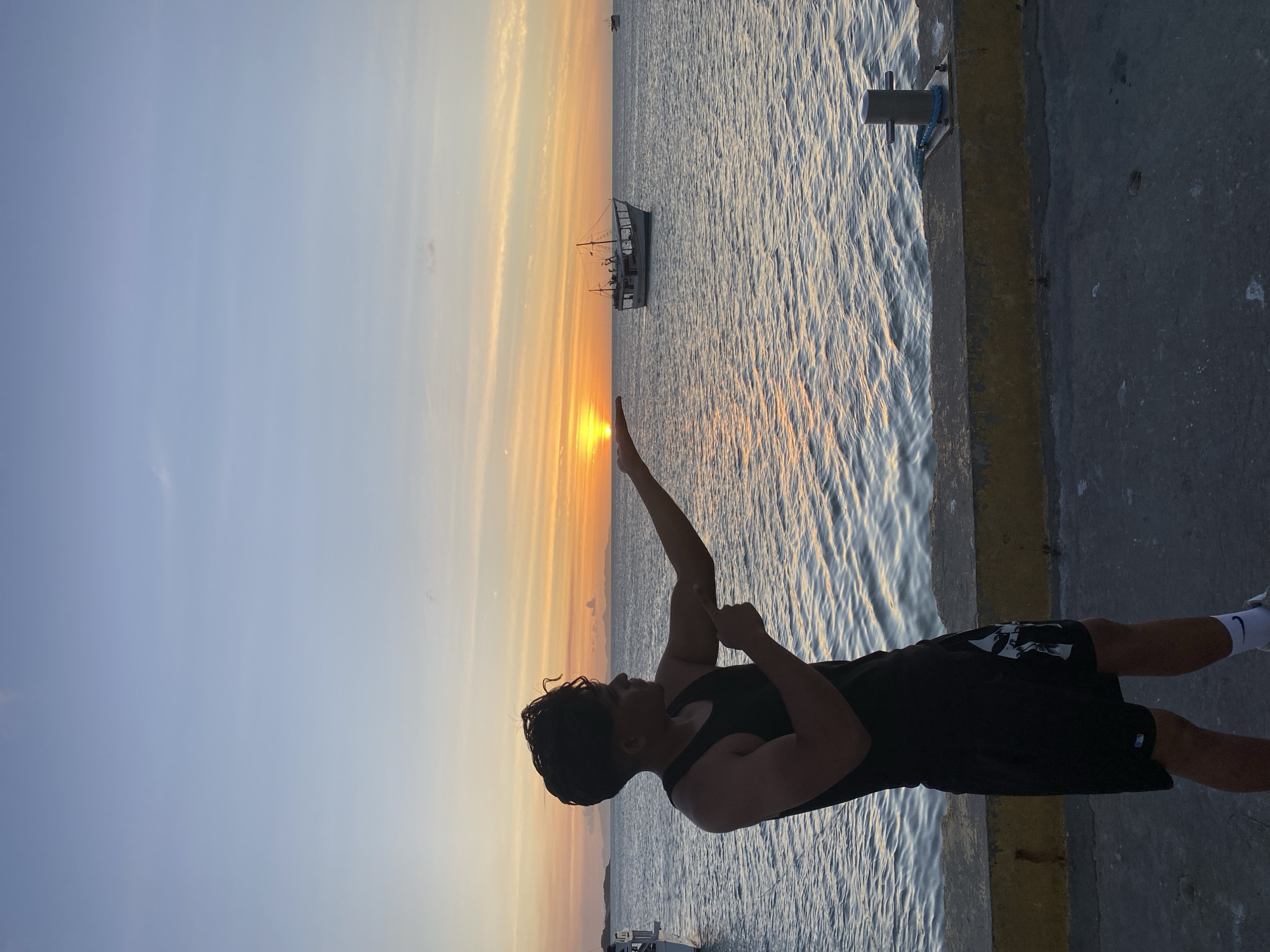 A man stands on a concrete pier at sunset in Juan Griego, Venezuela, gesturing towards the setting sun with his right arm. The man is silhouetted, wearing a dark tank top and shorts. A small boat is visible on the water in the distance, positioned between the man and the setting sun. The sky transitions from blue to orange as the sun sets on the horizon. The sea reflects the sunlight. A mooring bollard is fixed to the pier to the left of the man.