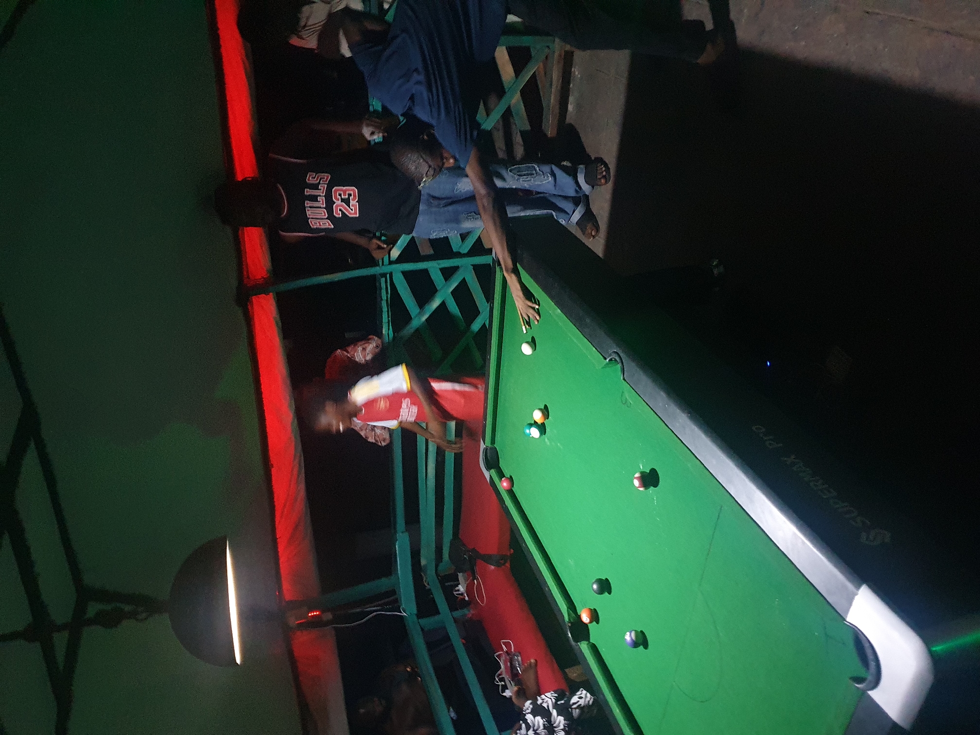 The image depicts a group of people playing pool at night in Lokoja, Nigeria. A man wearing a black Chicago Bulls jersey is standing near the pool table, observing a man in blue shirt who is positioned to take a shot at the white ball on the pool table. Another man in a red and white shirt and red pants stands to the side, also observing the game. The pool table, labeled "SUPERMAX PRO," is green, with multiple pool balls scattered across it. The area is illuminated by overhead lighting, which casts strong shadows. A structure with green-painted metal framing surrounds the area, with a section lined with red lights. The environment appears casual, suggesting a recreational setting or an outdoor entertainment area.