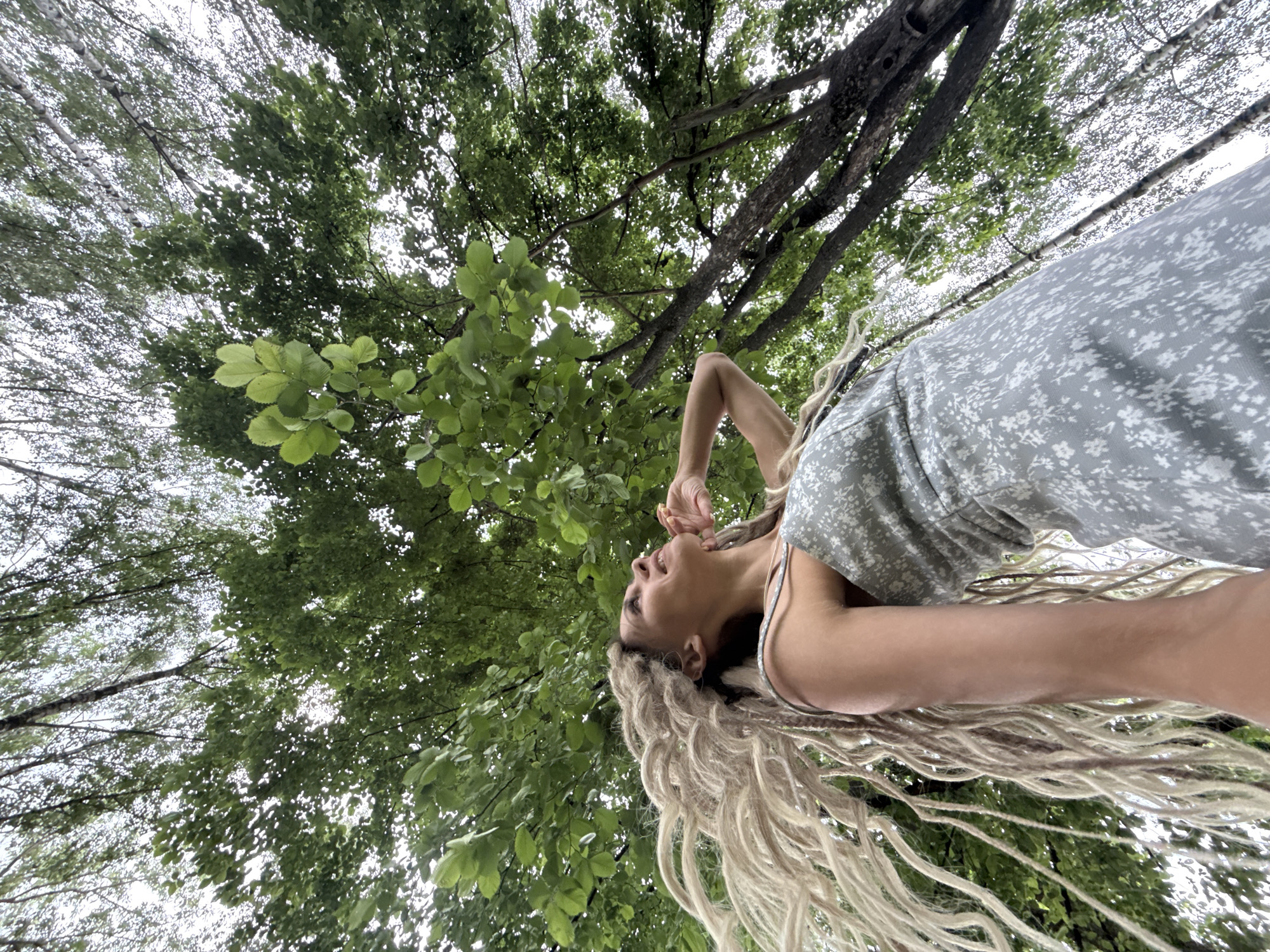 A low-angle shot looks up at a young woman with long blonde dreadlocks as she stands beneath a canopy of green trees. The woman is wearing a light gray dress with a subtle floral pattern. Her right arm is raised, with her hand near her face, as if she is enjoying the moment or contemplating something. Her head is tilted back, and her mouth is slightly open, suggesting she may be speaking, singing, or simply taking a deep breath.
The scene is outdoors, with the focus on the interplay of light and shadow filtering through the dense foliage of the trees. The leaves are a vibrant green, indicating a time of year when they are in full bloom, likely spring or summer. The sky is overcast, creating a diffused, soft light that evenly illuminates the scene. The overall mood is serene and natural, with the woman appearing to be at peace and connected with her surroundings. The image doesn't contain any visible text, and the specific location is not identifiable beyond being a wooded area. The time of day is likely daytime, given the natural light.