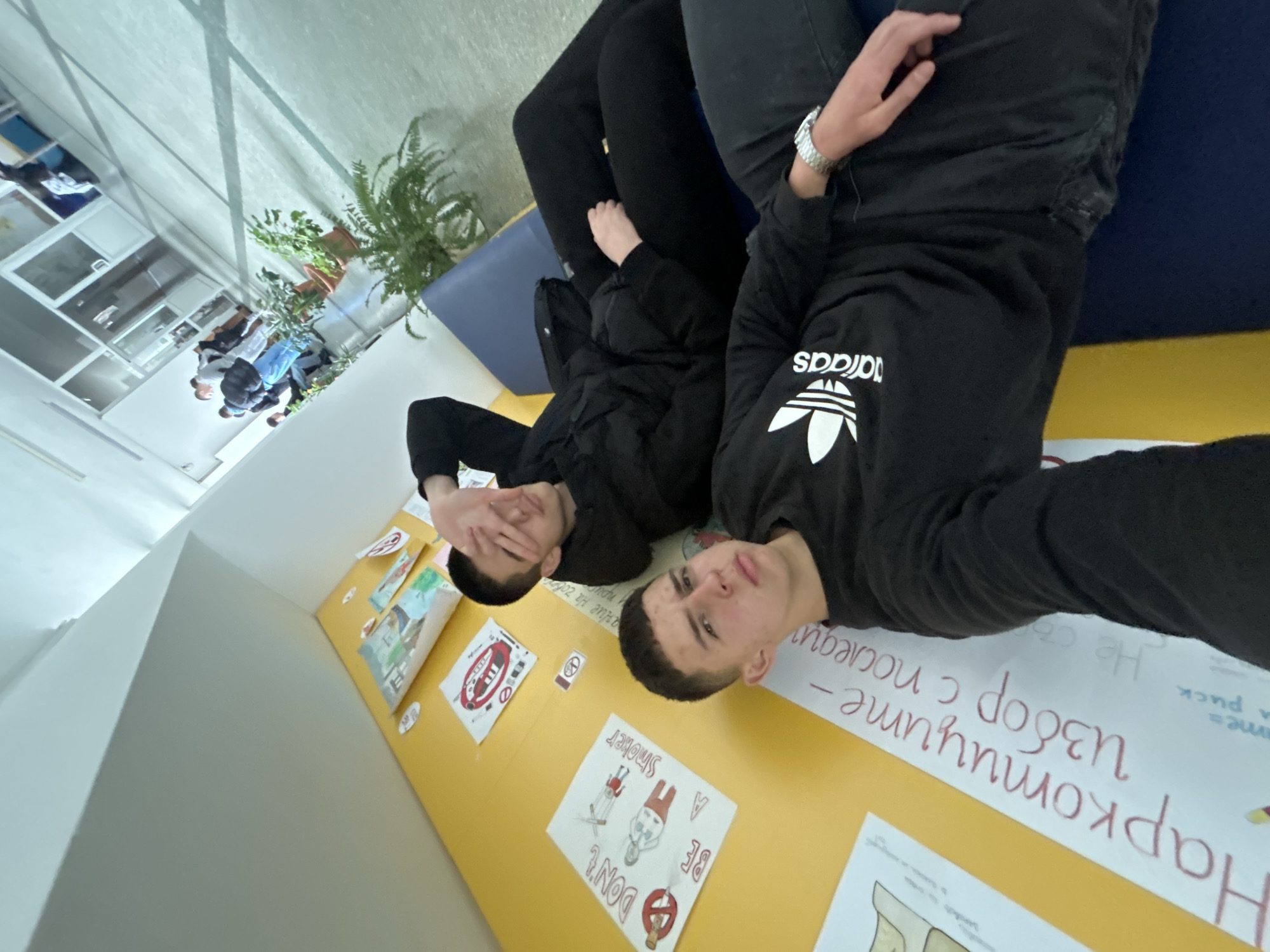 Two young men are lying on a yellow surface covered in posters, one wearing an Adidas sweatshirt. The posters appear to be public health messages related to smoking and narcotics. The location appears to be indoors near a window with plants and the street view visible. The text on the posters is Bulgarian, suggesting the location is in Bulgaria. Given this and the possible public health nature of the posters, it is likely that the location is an educational institution in Zlatograd, Bulgaria.