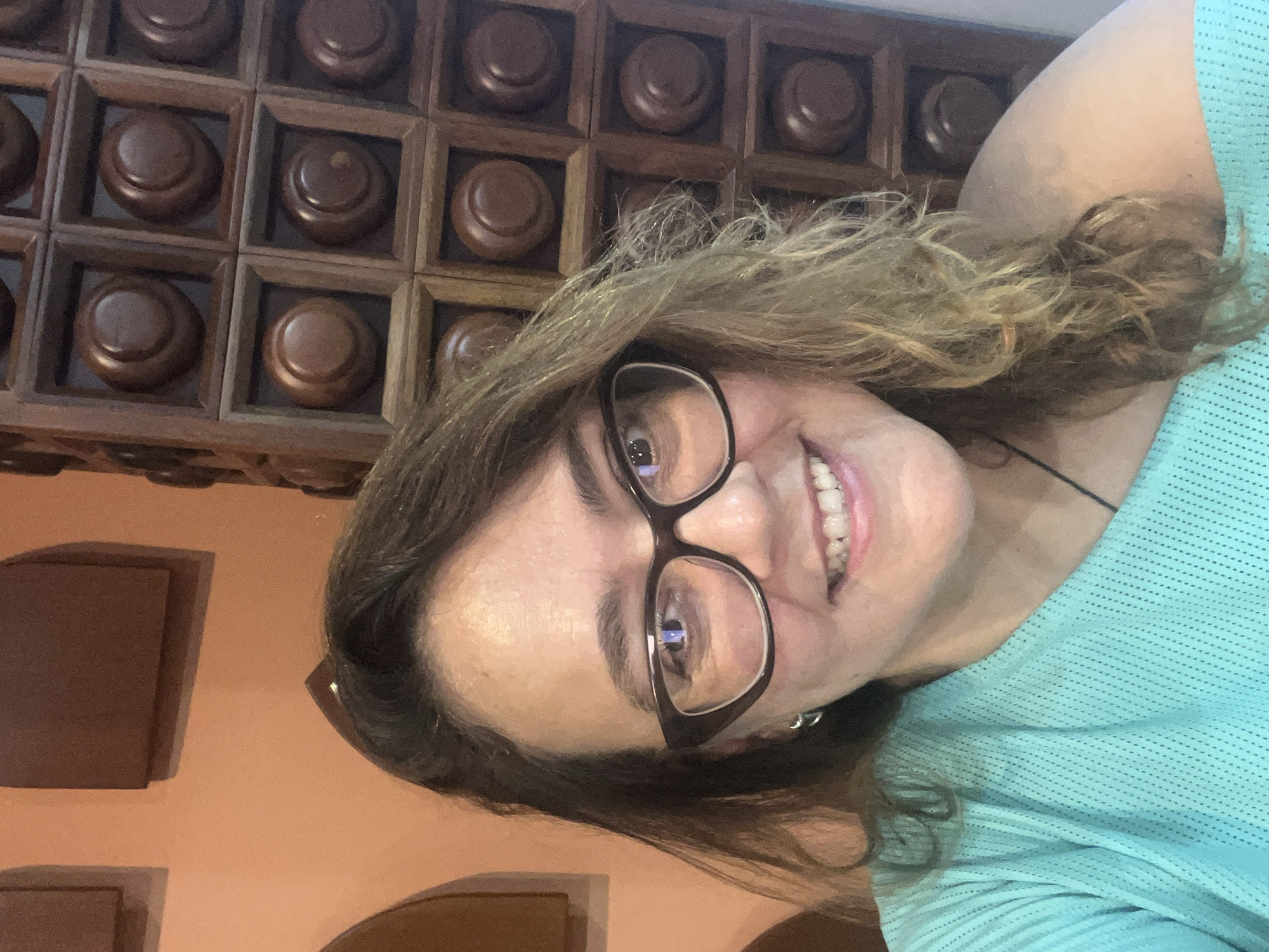 A medium-shot, front-facing photograph shows a woman wearing glasses and a teal shirt. The woman has brown hair and is smiling. She is positioned in front of a wooden shelf with multiple compartments, each containing a round wooden object. The background consists of a salmon-colored wall.