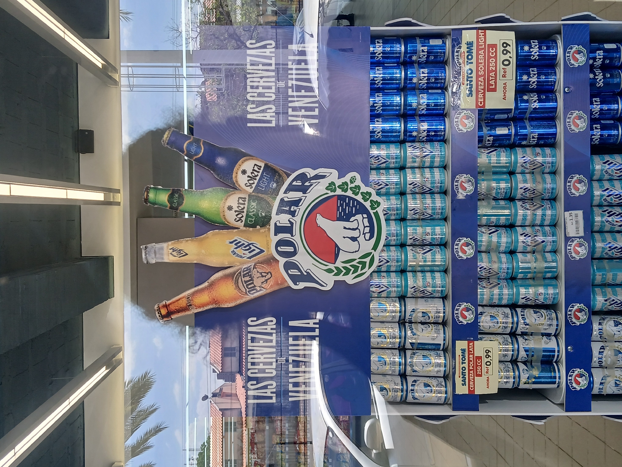The image displays a retail store interior, featuring a prominent beer display. The main subject is a large, vibrant promotional poster and an adjacent shelving unit filled with beer cans.
The poster showcases four different beer bottles: Solera Light (blue label), Solera Classic (green label), another Solera Light (yellow label, distinct from the first), and a Polar beer (amber bottle). Above these, the text "LAS CERVEZAS DE VENEZUELA" (The Beers of Venezuela) is boldly printed in white, indicating the origin of the products. A large "POLAR" logo with a polar bear icon is also visible.
To the right of the poster, a multi-tiered display unit holds numerous beer cans. The top shelves contain blue cans of Solera Light. Below these are light blue and white striped cans of Polar Light. The bottom shelves hold silver and gold cans of Polar beer.
Several price tags are attached to the display. One tag reads "SUPERMERCADO SANTO TOMÉ CERVEZA SOLERA LIGHT LATA 250 CC AHORA Ref 0,99," indicating a price of 0.99 for a 250cc Solera Light can. Another similar tag states "SANTO TOMÉ CERVEZA POLAR LATA 250 CC AHORA Ref 0,99" for Polar beer at the same price. A smaller, partially visible tag shows "1,35."
The setting is an indoor space with modern ceiling lights. A large window or glass door allows a reflection of the outdoors, showing clear skies, green palm trees, and buildings with red-tiled roofs, suggesting a warm, daytime environment in Ciudad Guayana, Venezuela. No people are visible, and the scene depicts a static product display.
