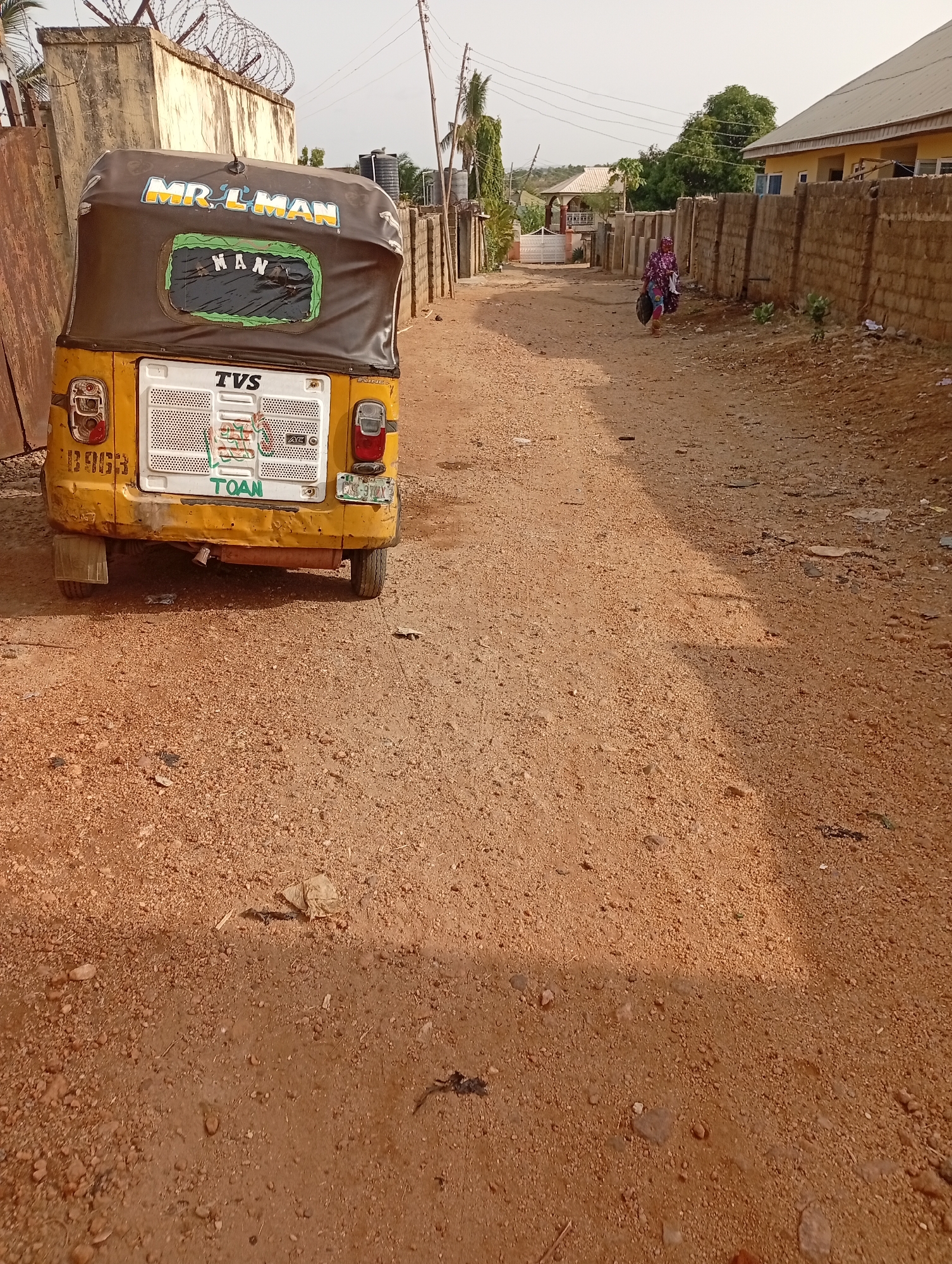 The scene is a dirt road in Adankolo, Nigeria. A yellow and black three-wheeled vehicle, labeled "Mr. L-Man" and "TVS," is parked on the left side of the road. A person is walking in the distance, towards the end of the road. Walls line both sides of the road. Barbed wire is visible atop one of the walls. Buildings are seen in the background.
